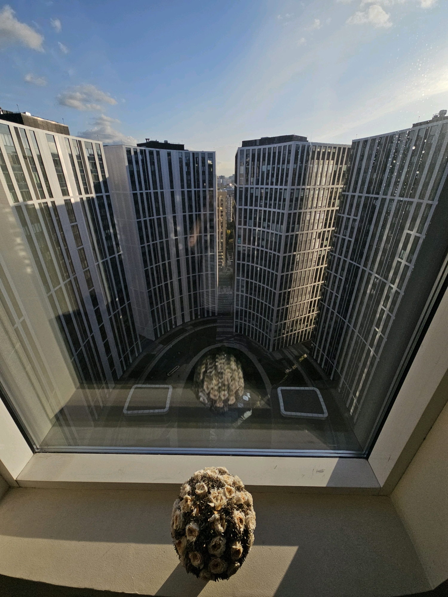 The image depicts a view through a window, looking out onto a modern architectural complex in Moscow, Russia. The foreground features a bouquet of dried flowers on a windowsill. Beyond the window, two large, curved buildings with repeating vertical elements dominate the scene. The sky above is a clear blue with scattered white clouds. In the courtyard between the buildings, a round floral arrangement is visible, possibly a sculpture or a decorative element. The architecture suggests a contemporary urban setting.
