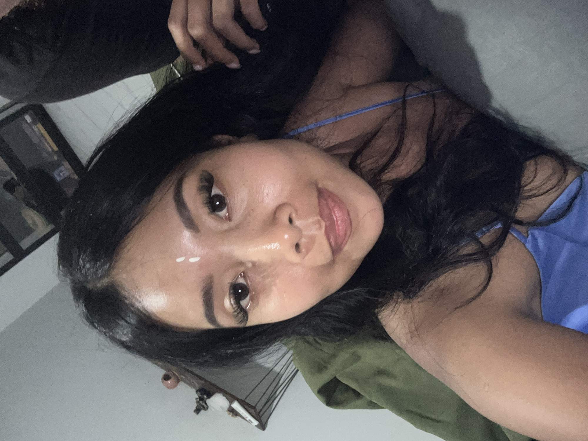 A selfie is taken from a low angle of a person with dark hair and light skin. The person is lying down, propped up on their left arm and shoulder, with their head tilted back. They are wearing a blue satin camisole with thin straps. The background is dark and out of focus, showing what appears to be a shelf with some objects on it and a dark piece of fabric in the upper right corner. The image has a shallow depth of field, with the person's face in sharp focus and the background blurred. There is a light patch on the person's forehead and a white mark on their nose. The overall impression is of a candid, casual moment captured at home. There is no indication of the location in the city of Sukawati, Indonesia.