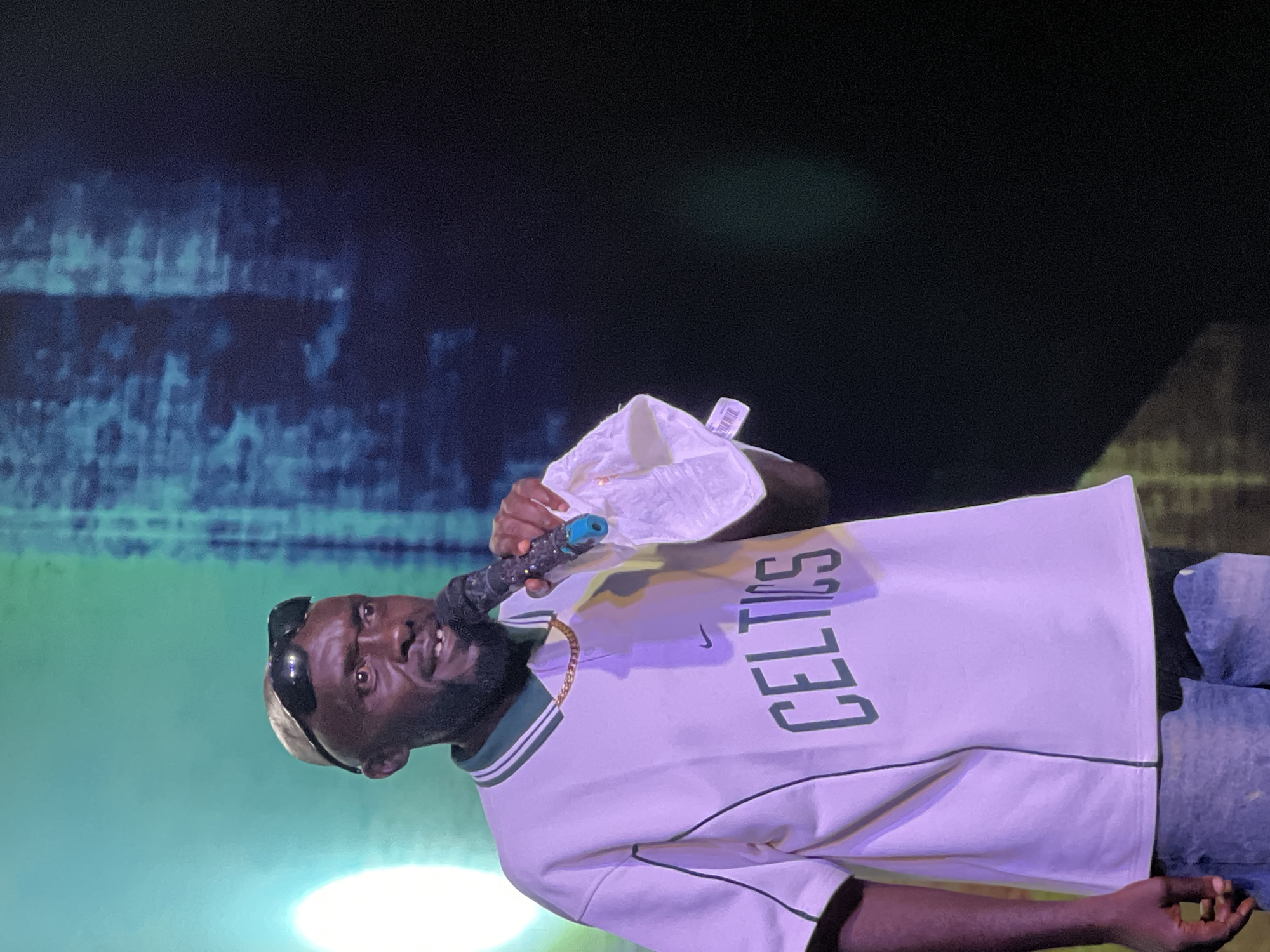 A man stands on a stage holding a microphone and a white cloth, speaking into the microphone. He is wearing a white polo shirt with green trim and the word "CELTICS" across the chest. He also has on a tan baseball cap and sunglasses. The background is dark with some blurred blue and green lights. The scene appears to be a performance or event. There is no information about the city of Gura Top, Nigeria in the image.