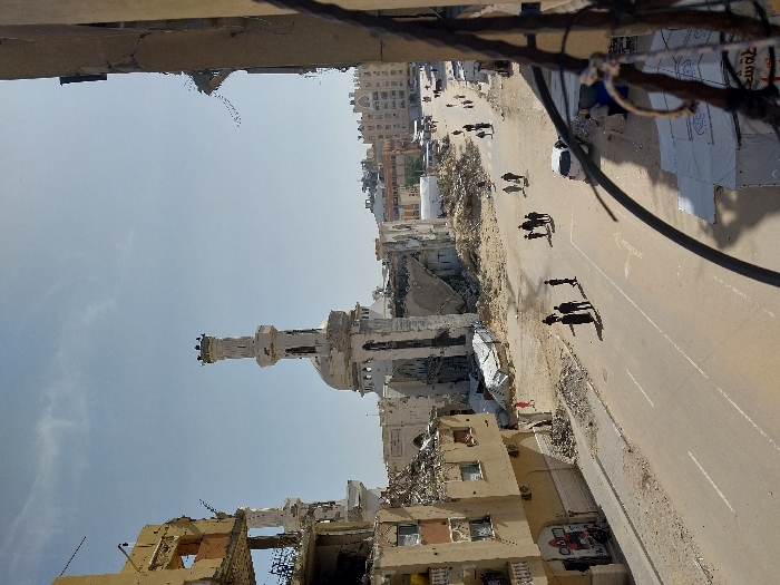 This image depicts a street scene in Khan Yunis, Palestinian Territory, characterized by extensive urban destruction under a clear sky with faint cirrus clouds. A central, partially destroyed mosque features a largely intact minaret with visible damage near its top, and a partially collapsed main structure. Surrounding buildings are heavily damaged, with multi-story structures reduced to rubble, exposing internal frameworks and missing walls. Piles of concrete, twisted rebar, and general debris litter the street sides and areas where buildings once stood. Approximately 15-20 individuals, appearing to be civilians, are walking along the paved street and scattered sidewalks, primarily in small groups or individually. A white passenger vehicle is parked on the right side of the road. Faint white road markings are visible on the asphalt. On the far right, a temporary white tent-like structure is set up. The photograph is taken from an elevated position, with damaged structural elements, including dark cables, visible in the immediate foreground on the right. Graffiti in red and white is visible on a lower section of a damaged building on the left.