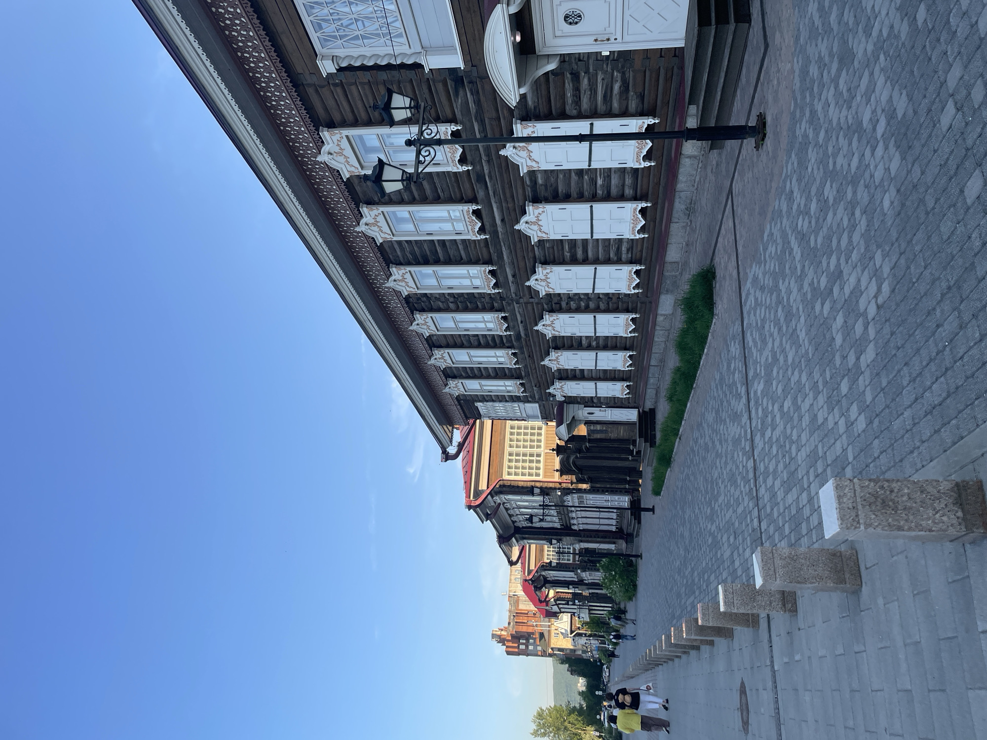 The image displays a clear daytime street scene in Krasnoyarsk, Russia, characterized by traditional architecture under a bright blue sky. On the left, a multi-story wooden building with intricate details dominates the foreground. Its dark wood facade is adorned with ornate white carved window frames and decorative elements along the eaves. Some windows are visible, while others are covered by closed white shutters. Black, decorative street lamps are mounted on the building's exterior.
To the right, a wide pedestrian pathway or street is paved with a pattern of grey and darker grey interlocking pavers, extending into the distance. A thin strip of green grass borders the building at ground level. Further along the paved area, a series of low, rectangular stone bollards or benches provides seating or visual demarcation.
In the background, other buildings of different styles are visible, suggesting an urban environment. Several people can be seen walking or standing along the paved area in the distance. Closer to the foreground, two individuals are more clearly visible on the right: one wearing a yellow top and dark pants, and another in a white top and dark pants, seemingly strolling casually.
The weather is clear and sunny, indicated by the bright blue sky with only faint clouds and the natural lighting of the scene, suggesting either late morning or early afternoon. The overall impression is one of a peaceful, well-maintained urban area featuring historical architectural elements. There is no visible text in the image.