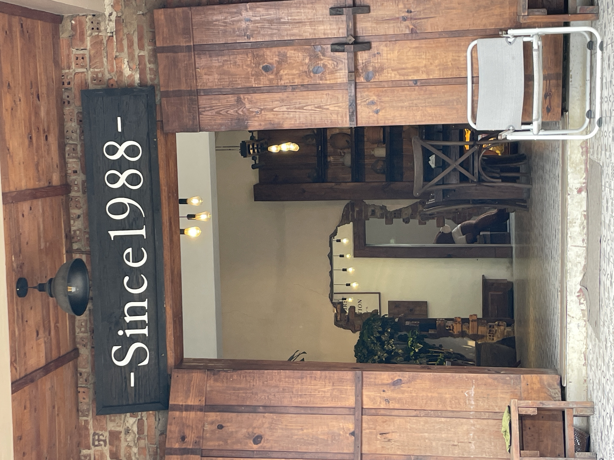 The image shows the entrance to a cafe or restaurant in Phường Xuân Hương - Đà Lạt, Vietnam. A dark wooden sign with white text reads "-Since 1988-". The entrance is framed by two large, rustic wooden doors, one of which is slightly ajar, revealing the interior. Inside, there are tables, chairs, shelving with decorative items, and plants. Several pendant lights hang from the ceiling. Outside the entrance, a light-colored folding chair sits on the tiled ground. The building's exterior features exposed brickwork and wooden beams supporting a roof. The overall aesthetic suggests a vintage or rustic theme. No people are clearly visible within the frame of the main entrance.