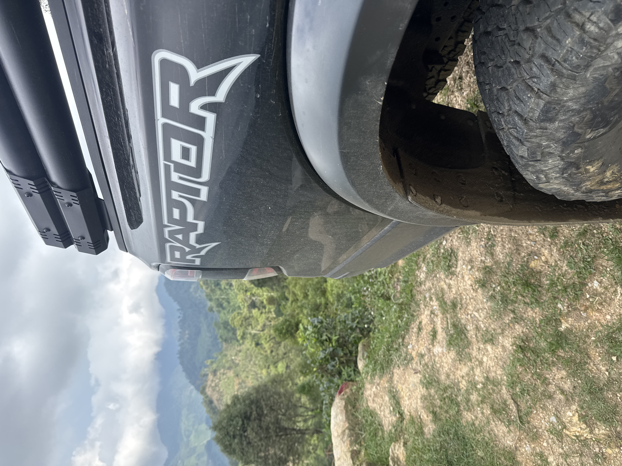 The image shows the rear of a black Ford Raptor truck parked on an unpaved surface overlooking a valley in Suối Giàng, Vietnam. The "RAPTOR" emblem is visible on the rear quarter panel. The truck's tire is visible on the right side. The landscape is characterized by lush greenery and mountain ranges in the distance under an overcast sky. The ground immediately around the truck consists of dirt, grass, and small rocks.