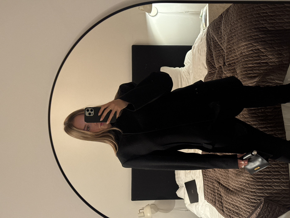 A selfie captures a woman in a black coat, reflected in a round mirror. She holds her phone up, partially obscuring her face. Behind her, a bed with a brown quilted comforter and a black headboard are visible. A bedside lamp and a laptop rest on the bed. The room appears to be well-lit with a warm tone. She is holding a small black handbag.