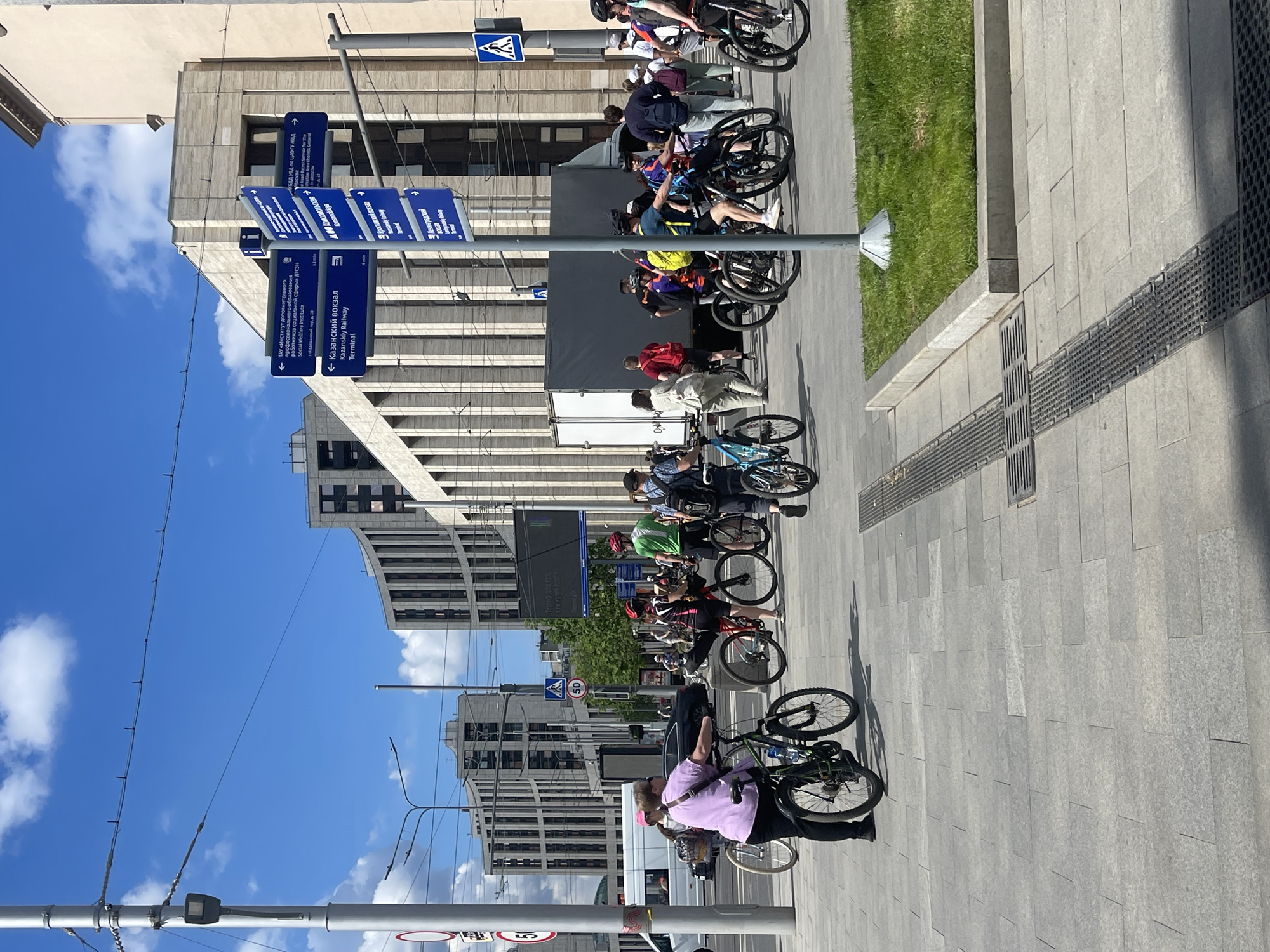 The image shows a group of cyclists stopped on a wide sidewalk in Moscow, Russia. A sign directs toward Kazanskiy Railway Terminal. Some cyclists are on their bikes, while others are standing beside them. A delivery truck is parked nearby. Buildings are in the background, along with blue skies and some clouds. The day is sunny. A sign showing the route to the train station is near the cyclists.