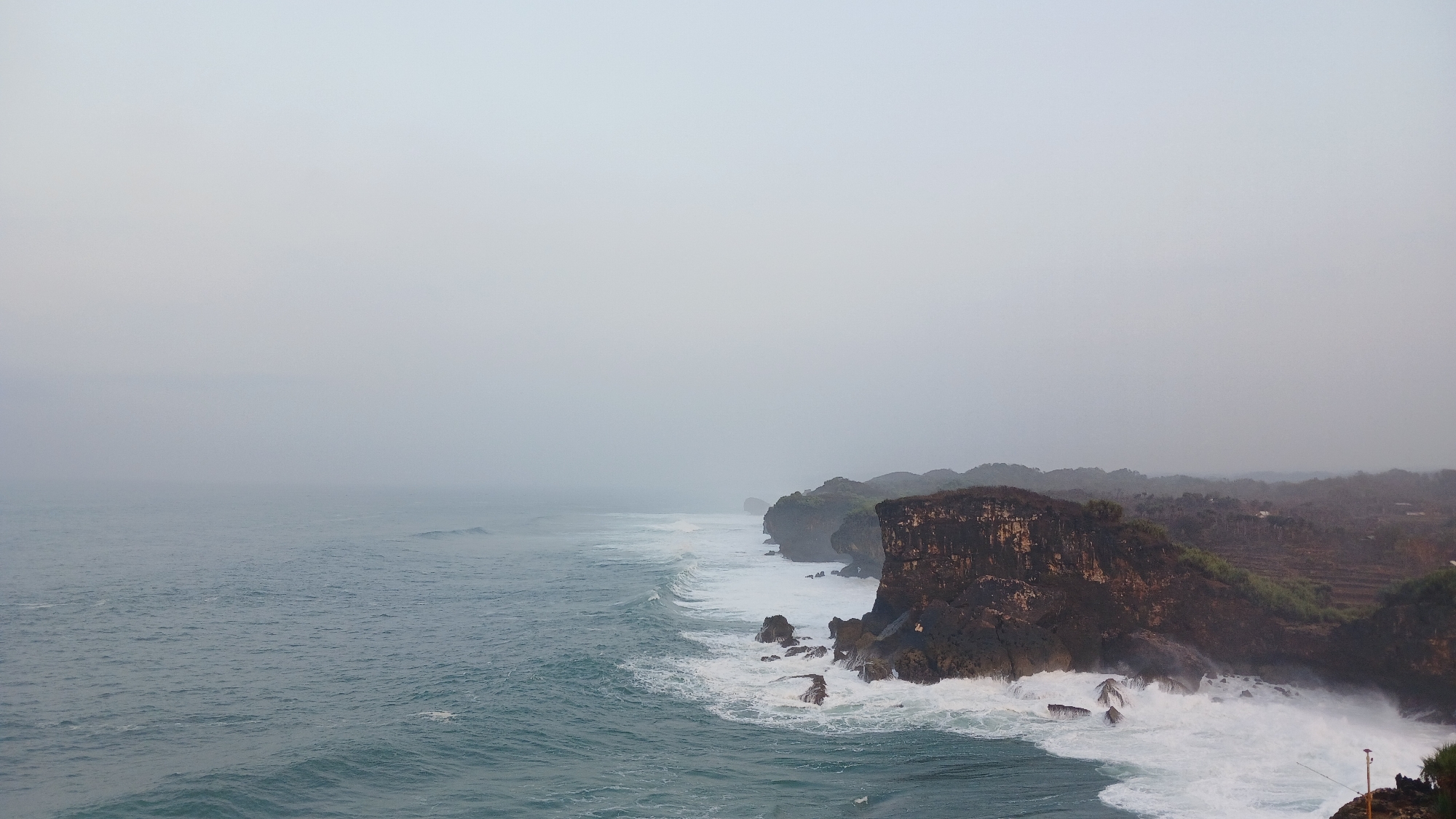 The image captures a scenic view of the coast in Tanjungsari, Indonesia. The scene is dominated by a rugged coastline with steep, rocky cliffs that plunge into the turquoise ocean. Waves crash against the shore, creating white foam and spray, particularly around the base of the cliffs. The water appears choppy, with visible ripples and waves stretching towards the horizon.
The sky is overcast and hazy, suggesting a cloudy or foggy day, which lends a soft, diffused light to the scene. The distant landscape behind the cliffs is covered in lush green vegetation and appears to be terraced, indicating agricultural activity or natural undulating hills. There is no sign of human presence or man-made structures in the immediate vicinity of the cliffs, reinforcing the natural and somewhat remote feel of the location. The overall impression is one of natural beauty and the power of the ocean.