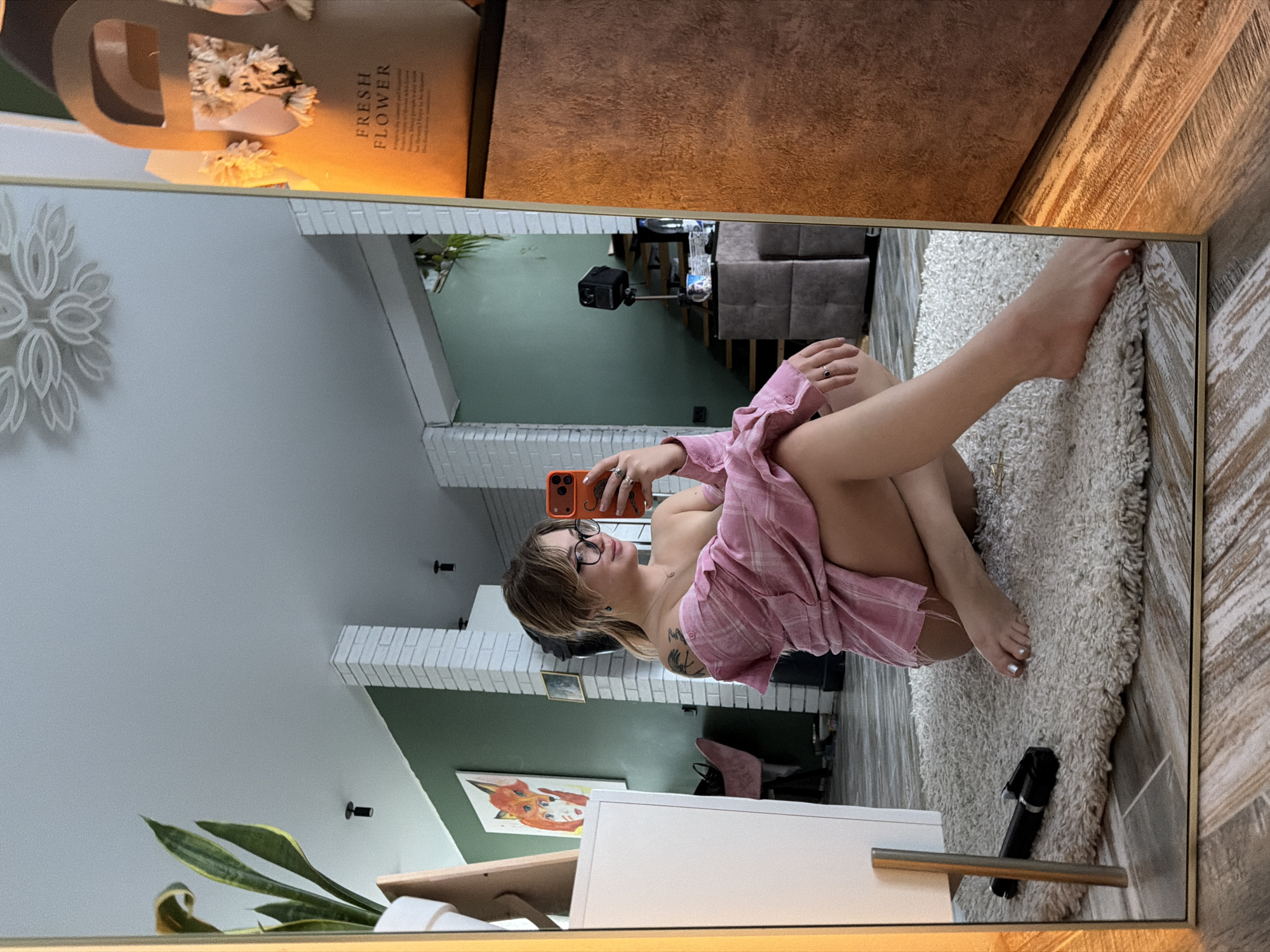 Reflected in a large mirror, a young woman takes a selfie in an indoor setting, likely a home in Большая Шильна, Russia. She is seated on a shaggy white rug, leaning back with her left leg extended and her right leg bent. She holds an orange smartphone to capture her reflection, wearing a pink plaid shirt and glasses. A tattoo is visible on her left arm, and her fingernails and toenails are painted. She appears to be smiling or smirking, conveying a relaxed and confident mood.
The room, as seen in the reflection, features walls painted in a light grey and a darker green shade, accented by white brick-style detailing. On the right, wood-look paneling or flooring is visible. In the upper left corner of the mirror, a white, intricately designed wall decoration is mounted. In the top section of the image, above the mirror, a bouquet of white flowers in a brown paper bag with the text "FRESH FLOWER" is visible.
Other objects in the reflection include a painting of a fox on the green wall, a black selfie stick lying on the rug near the woman's feet, and a camera on a tripod further in the background. A houseplant with long, variegated leaves is situated near a white dresser or cabinet at the bottom of the frame. The lighting suggests indoor artificial light, contributing to a bright interior scene.