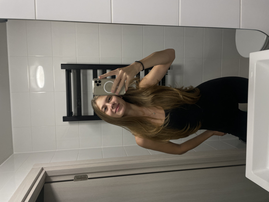 A young woman takes a mirror selfie in a bathroom. She is wearing a black tank top and leggings. She holds a phone up with her right hand. The bathroom has white tile walls, a gray door, a black towel rack, and a white sink. The woman has long brown hair. She is smiling slightly at the camera.