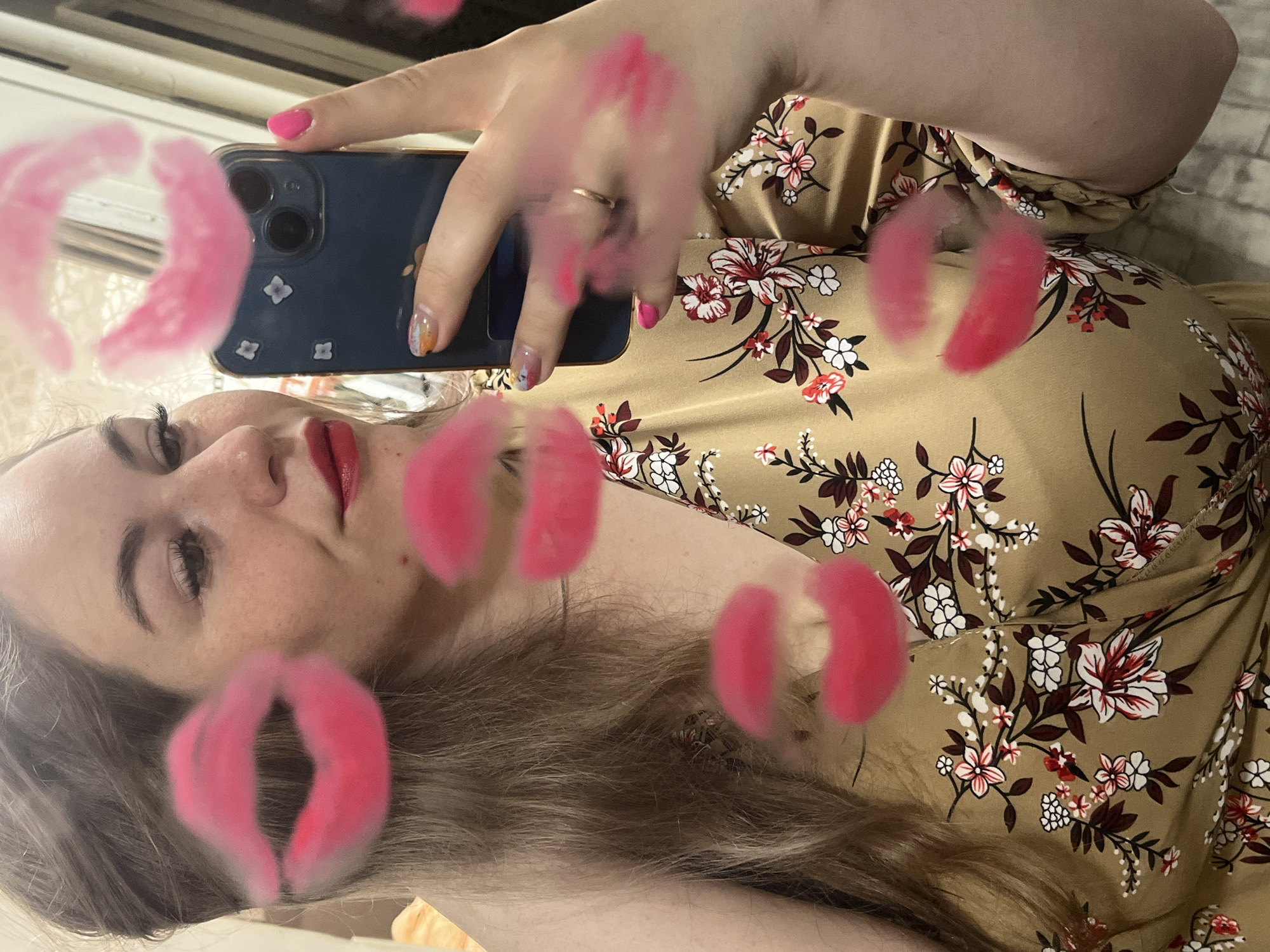 A woman is taking a selfie in a mirror. She is holding a dark blue phone with flower stickers on the back. She is wearing a floral print dress with a tan background. Her fingernails are painted, one in a solid pink and another with multicolored designs. She is wearing a ring on her finger. She is wearing red lipstick. There are digital pink lip graphics added to the image. The location is unknown and no evidence suggests it's in Chaikovsky, Russia.