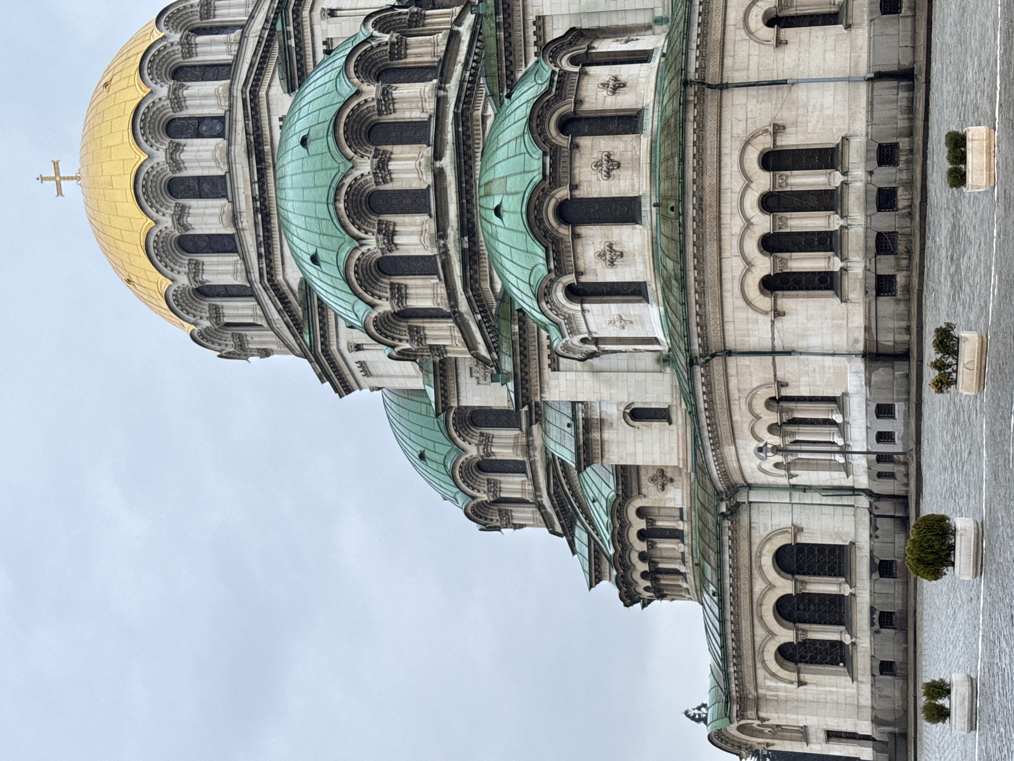 The image shows a portion of the St. Alexander Nevsky Cathedral in Sofia, Bulgaria, viewed from a low angle. The building's architecture features multiple domes, one gilded with a cross at its apex, and several others covered in a light-green material. The exterior walls are primarily white or light gray, with ornate arched windows and decorative elements. The adjacent ground is paved with cobblestones and decorated with planters, some containing small shrubbery. The sky is overcast, contributing to a subdued lighting of the scene.