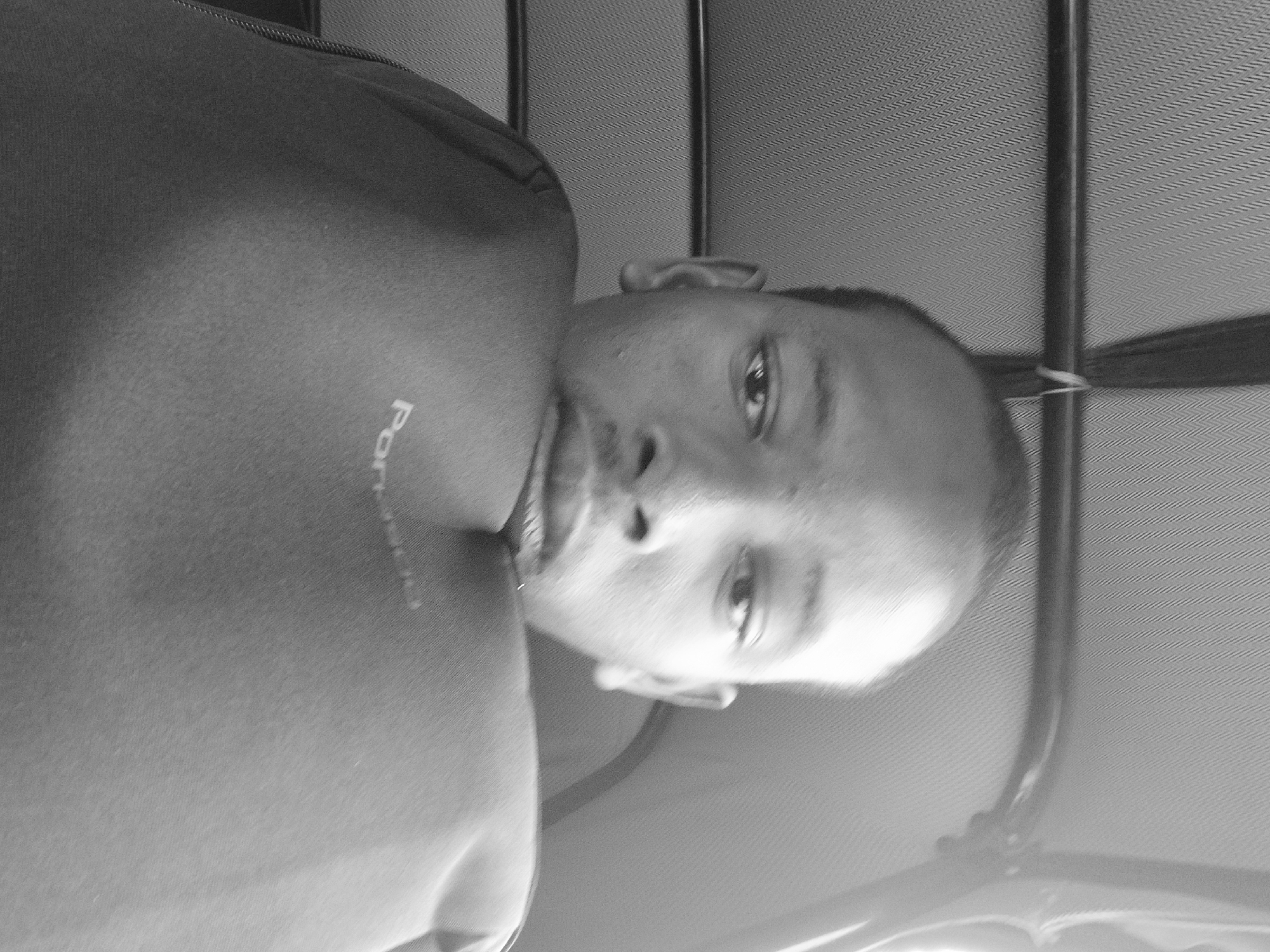 The image is a grayscale, low-angle, close-up selfie of a young person lying down. The person's head is tilted back, and their gaze is directed upwards and to the right. Their eyes are open, and their mouth is slightly parted, with a hint of a smile. A faint text, likely a logo or brand name, is visible on the person's shirt, appearing to read "POR". The background consists of a textured surface, possibly the ceiling of a vehicle, with a dark vertical bar and a thin strap running across it. The lighting suggests it could be daytime, possibly indoors or in shaded conditions. The image captures a candid moment, conveying a sense of casualness. The setting appears to be indoors, given the presence of the textured ceiling and the vertical support structure, which could be part of a car interior or similar enclosed space. The person's relaxed posture and direct gaze suggest they are comfortable and aware of being photographed. No specific weather conditions are discernible. The overall mood is informal and personal.
