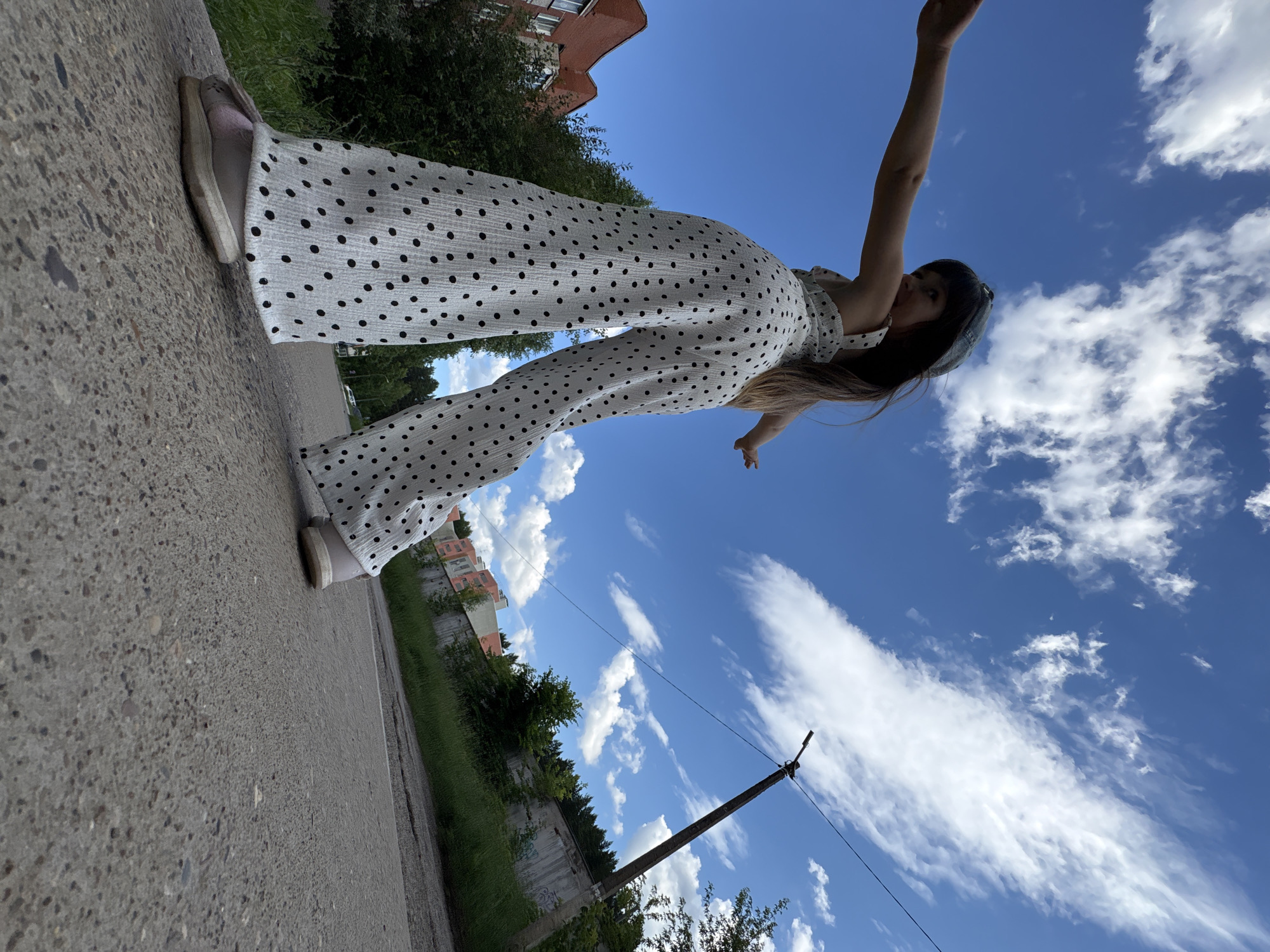 The image shows a person wearing a white polka dot jumpsuit and light-colored shoes, standing on a paved surface. The person is positioned at an angle, with their arm extended upwards and their body tilted. The background consists of a bright blue sky with scattered white clouds, lush green grass, trees, and some buildings in the distance. A utility pole is also visible. The scene appears to be outdoors on a sunny day, possibly in a semi-urban or suburban area. The perspective of the camera, angled upwards towards the sky, gives a sense of height and openness. There is no visible text in the image. The overall mood seems light and possibly playful due to the person's pose. The location context of Chaikovsky, Russia, suggests this could be a view of a typical Russian summer day.