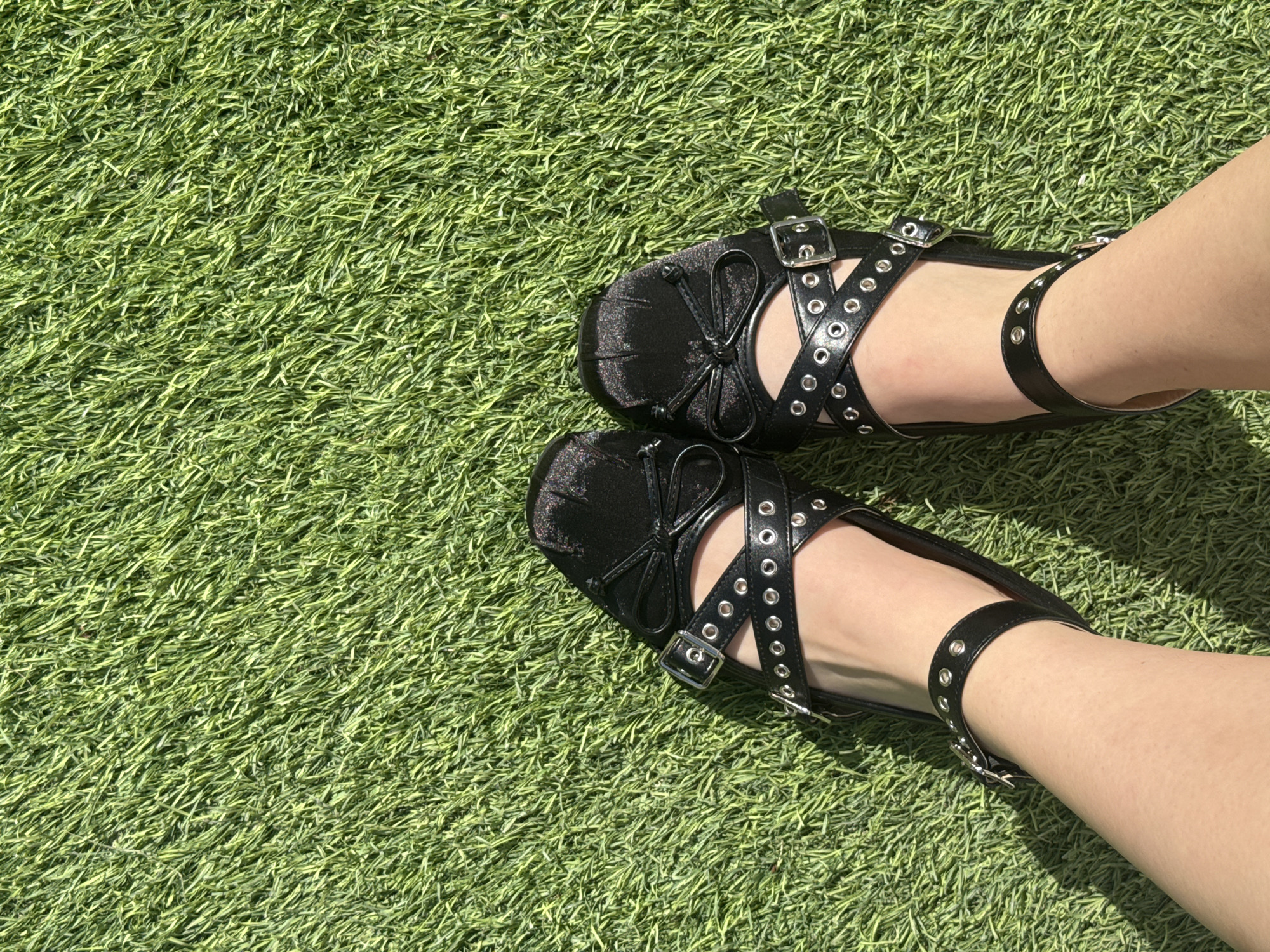 The image shows a pair of legs wearing black, studded Mary Jane shoes on a green, artificial turf surface. The shoes have a bow detail on the toe and a buckled ankle strap. The legs are pale and photographed from above. The background is solely the artificial turf. There is no evidence in the image to indicate this is Santa Clara Ocoyucan, Mexico.