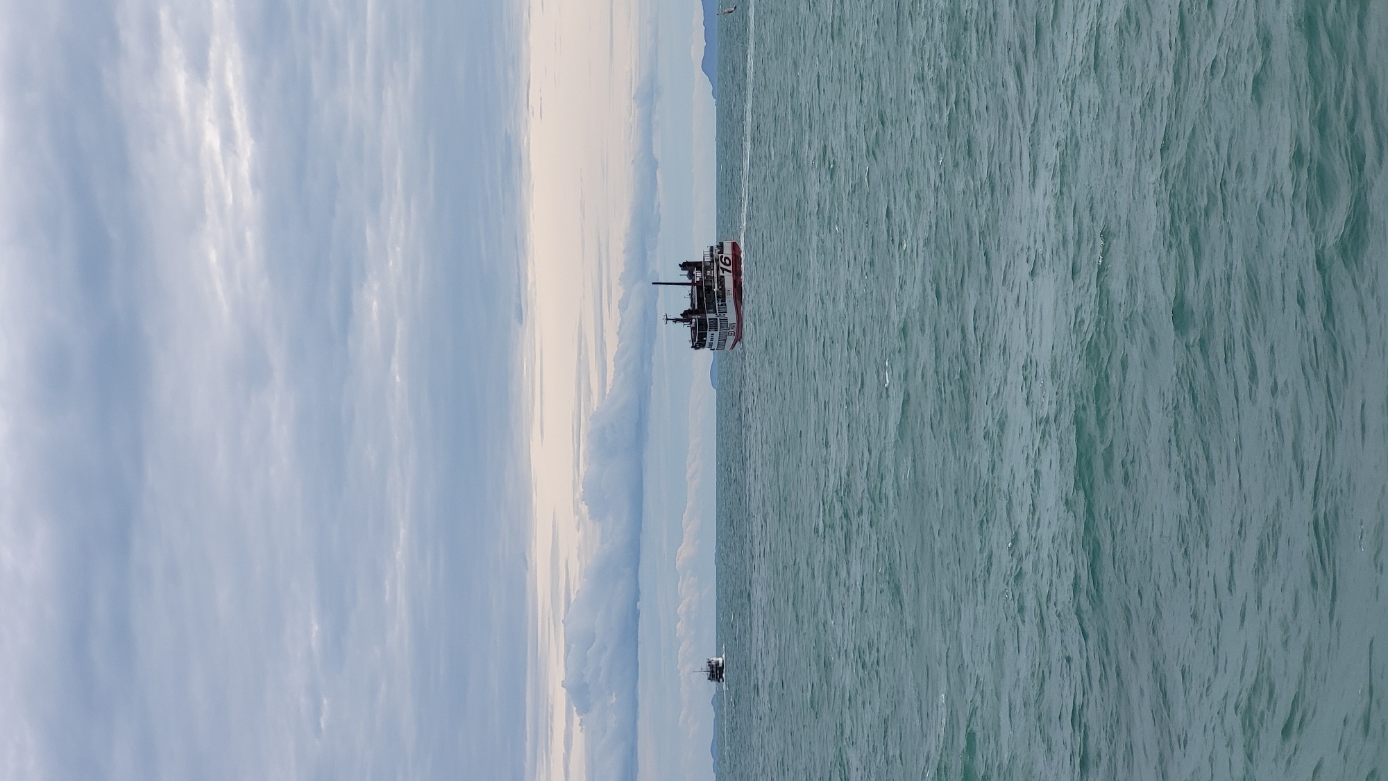 The image depicts a body of water with two ferries in the distance. The water is choppy and a greenish-blue color. The sky is overcast with grey clouds. On the right side of the image, a larger ferry is visible, painted white with red accents and the number "16" on its stern. On the left side, a smaller ferry is visible, also appearing to be white. In the far distance, there are faint outlines of landmasses, likely islands. The scene suggests a maritime environment, possibly a ferry route. This type of scenery is common in coastal areas of Ko Pha-ngan District, Thailand, where ferry transport is a primary mode of travel between islands and the mainland. There are no people clearly visible in the image.