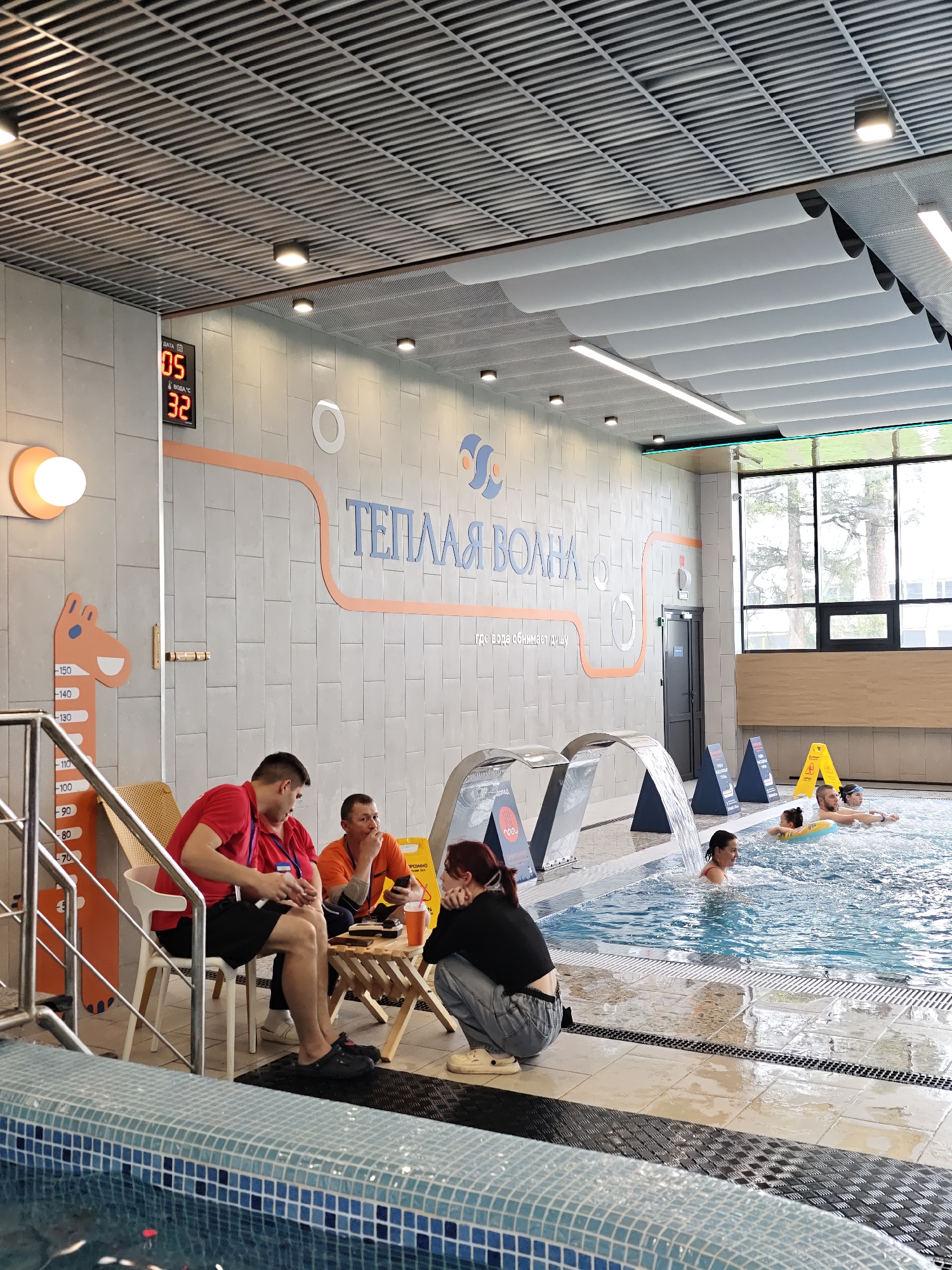 The image shows the interior of a swimming pool facility, likely located in Votkinsk, Russia, based on the Cyrillic text "ТЕПЛАЯ ВОЛНА" on the wall, which translates to "Warm Wave." A digital display shows the date as the 5th and the water temperature as 32 degrees Celsius.
Along the poolside, three individuals are seated at a small table. One man is wearing a red shirt, another an orange shirt, and a woman is kneeling in jeans and a black shirt. It seems they are taking a break or possibly eating.
In the pool, several people are swimming and enjoying the water. There are built-in water features resembling small waterfalls. Warning signs are placed at the edge of the pool.
The architecture includes a modern ceiling with visible infrastructure, tiled walls, and large windows providing natural light. The general environment appears clean and well-maintained.