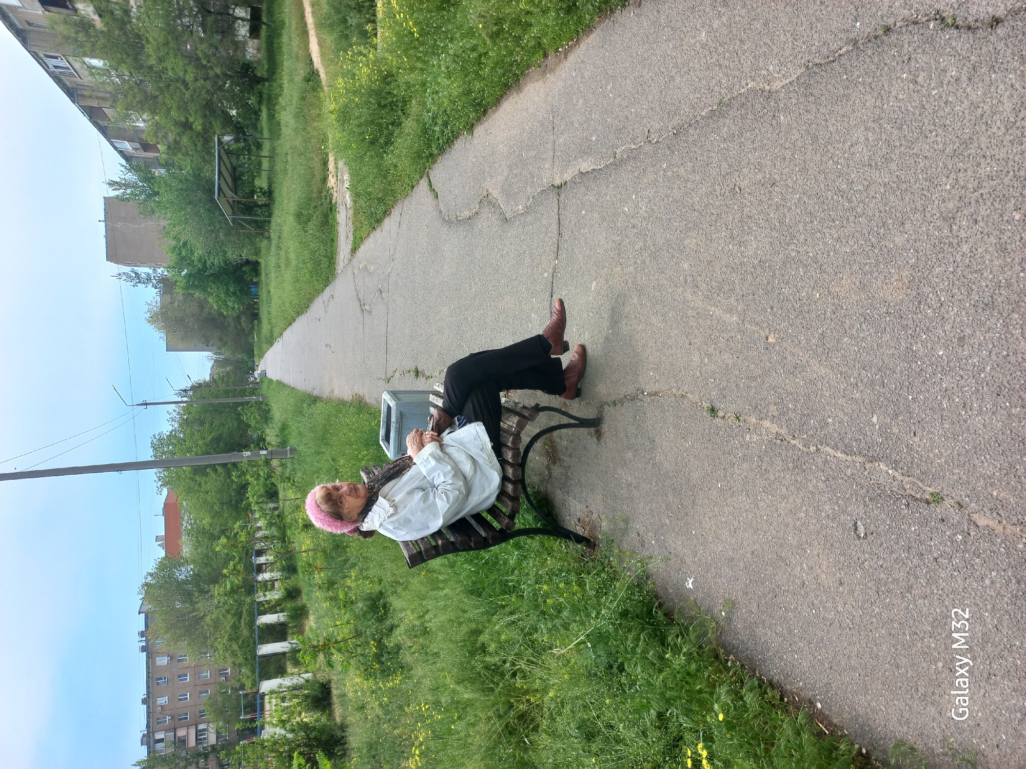 An elderly woman wearing a pink headscarf, a light-colored jacket, and dark pants sits on a park bench alongside a paved path. She is facing toward the camera, appearing relaxed. Her feet, clad in brown shoes, rest on the bench. The bench is positioned next to a trash can. The pathway extends into the distance, flanked by green grass and vegetation. Buildings are visible in the background on both sides of the path. The camera model, a Galaxy M32, is written in the bottom right corner of the photo. The buildings and typical green spaces suggest a location in a residential area, consistent with public park locations in Volzhsky, Russia.