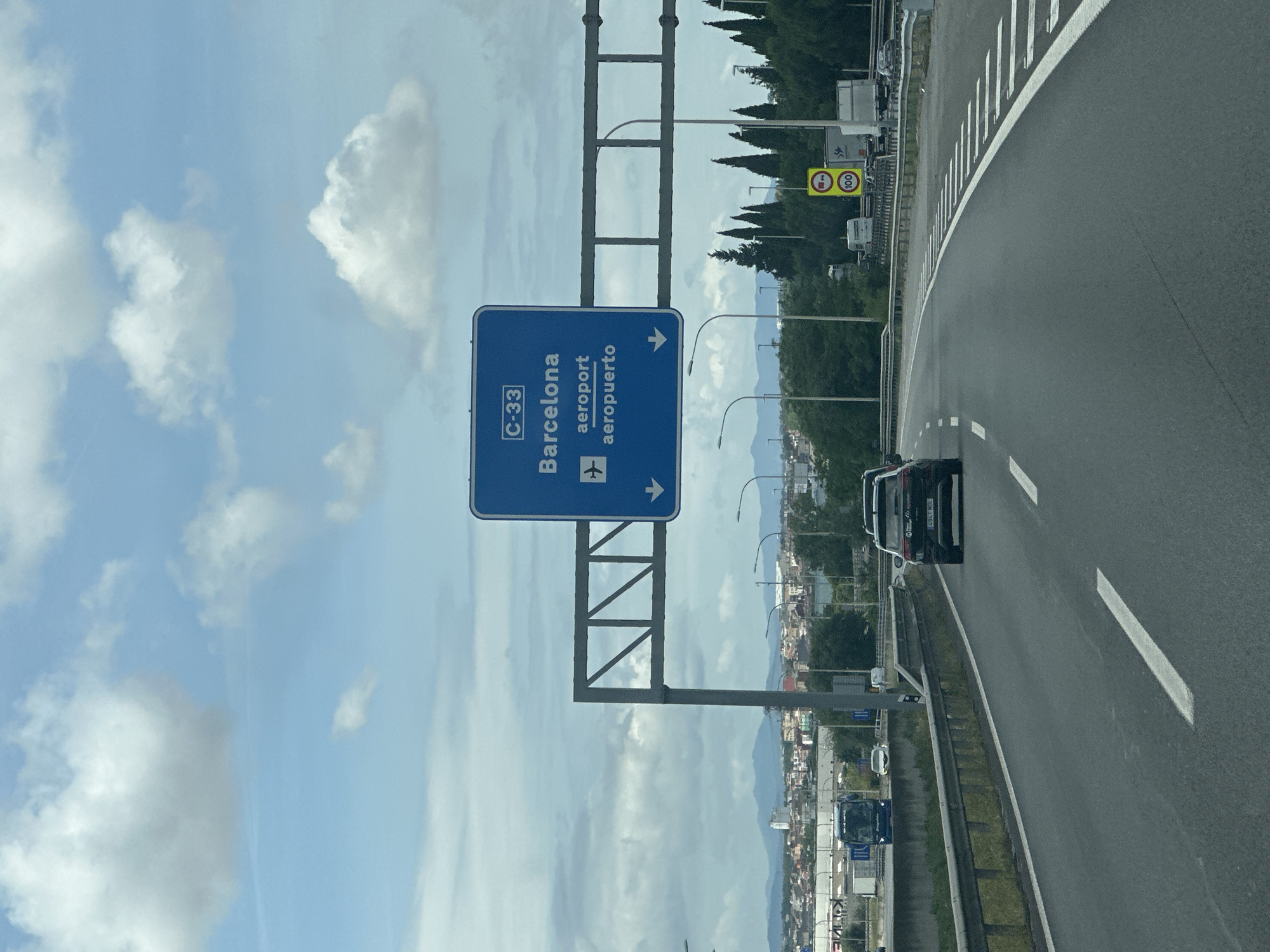 The image captures a daytime scene on a multi-lane highway, viewed from a moving vehicle. A large, blue overhead highway sign is prominently featured in the midground, indicating directions. The sign displays "C-33" at the top, followed by "Barcelona" and "aeroport" (Catalan for airport) with an airplane icon, and below that "aeropuerto" (Spanish for airport). White arrows on the sign point towards the right, indicating the direction for these destinations. The sign is supported by a gray metal gantry structure spanning across the road.
To the right of the highway, further in the distance, a yellow rectangular sign with a red border displays the number "100" and a truck pictogram, likely indicating a speed limit for trucks or specific lane usage. The highway itself is asphalt with clear white lane markings and a white solid line on the shoulder. It curves gently to the right. A black car is visible in the rightmost lane.
Along the right side of the highway, there's a metal guardrail, followed by a strip of grass and a line of tall conifer trees. Beyond these trees, a landscape of low mountains or hills is visible under a partly cloudy sky. More vehicles, including trucks and a blue bus, can be seen on the highway and what appears to be a lower road structure in the background. The sky above is bright blue with scattered white clouds. The setting is a clear day, indicating good visibility. Based on the visible text and context, the location is a highway in Spain, likely near Montmeló, leading towards Barcelona and its airport.