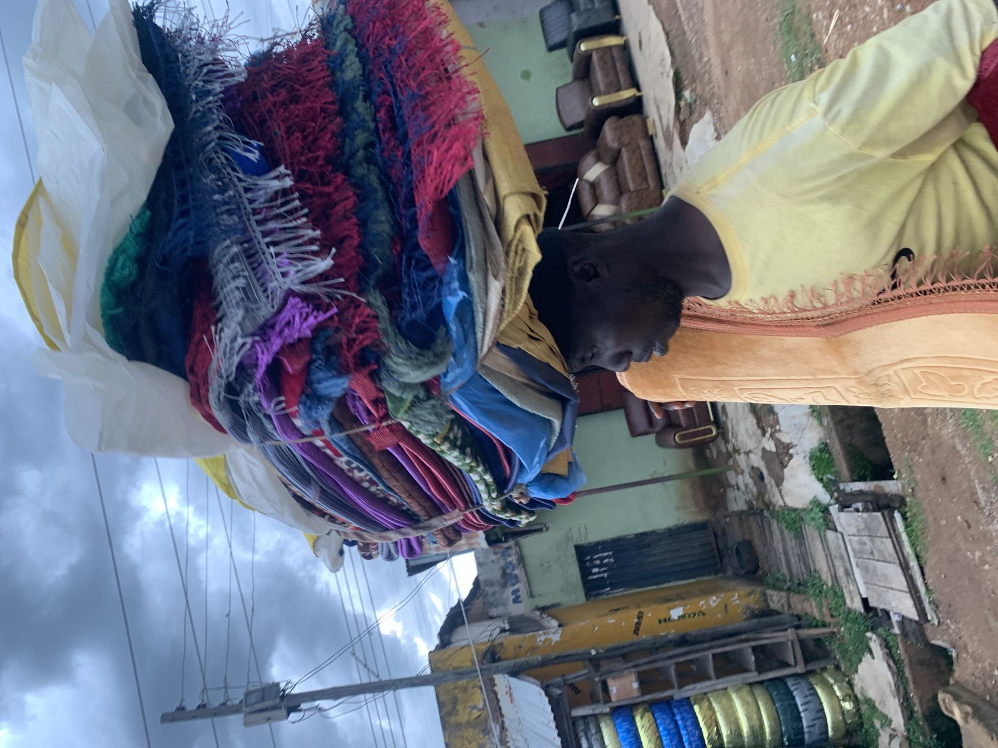 A person is seen carrying a large bundle of colorful textiles on their head. The textiles are predominantly in shades of red, blue, green, purple, and white, with some appearing to be fringed or tasseled. The person is wearing a light yellow robe with decorative trim. The scene appears to be outdoors, with a cloudy sky overhead. In the background, there are buildings with green walls and some furniture visible. There is also a yellow structure with shelves holding what appear to be tires. The ground is dirt or gravel. The image is oriented vertically. The overall impression is that of a market or street vendor scene, likely in Jos, Nigeria, given the context. The time of day is difficult to ascertain precisely due to the clouds, but it appears to be daytime.