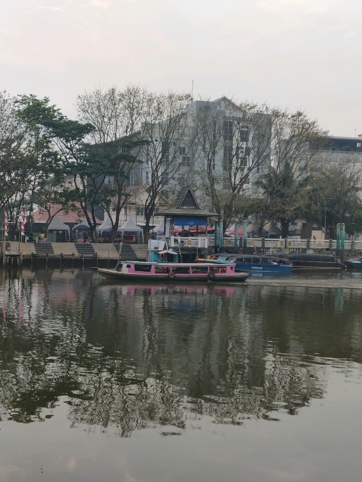 The image depicts a tranquil scene along the Martapura River in Banjarmasin, Indonesia. In the foreground, the murky green water reflects the surrounding environment, creating a textured, abstract pattern. A narrow, pink and green boat is positioned mid-river, its smooth surface contrasting with the rippling water. Behind the boat, a larger blue vessel with "BUNG LOW" printed on its side moves slowly. The riverbank is lined with trees, some bare of leaves, interspersed with buildings and small structures with pitched roofs. A white fence with blue posts runs along the embankment, and steps lead up to a raised walkway where several white tents and small market stalls are visible. In the background, a multi-story building with numerous windows stands prominently. The sky is overcast and a pale grey, indicating either early morning or late afternoon. The overall atmosphere is calm and still, with no prominent human activity.