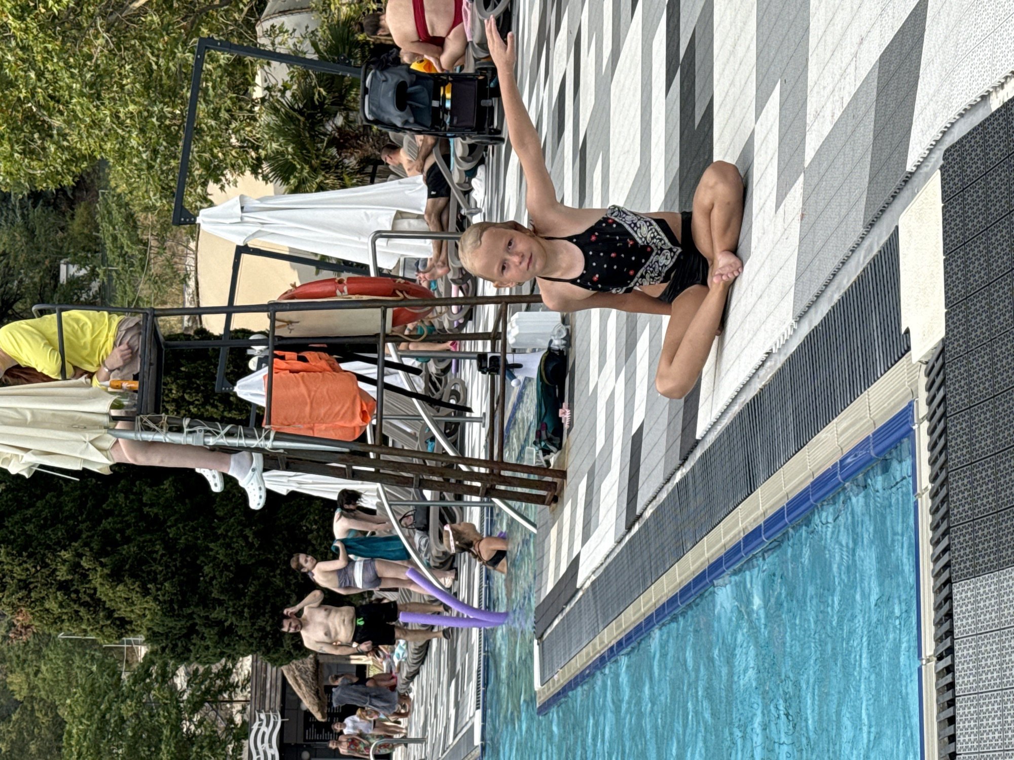 The image depicts a poolside scene with several people. In the foreground, a young girl in a black and white patterned swimsuit is sitting on the tiled ground next to a swimming pool, with her right arm raised. The pool has clear blue water and a tiled edge. The ground around the pool is paved with a black and white checkered pattern.
In the background, there are various objects and people. A metal structure with an orange life preserver and a white umbrella is visible. Several adults are seen in swim attire, some standing and some sitting. One person in a bright yellow shirt is lying down. Trees and foliage form the backdrop. The scene appears to be outdoors, likely during the day, with bright lighting suggesting fair weather. The overall atmosphere seems relaxed and leisurely, typical of a pool environment. There is no visible text in the image.