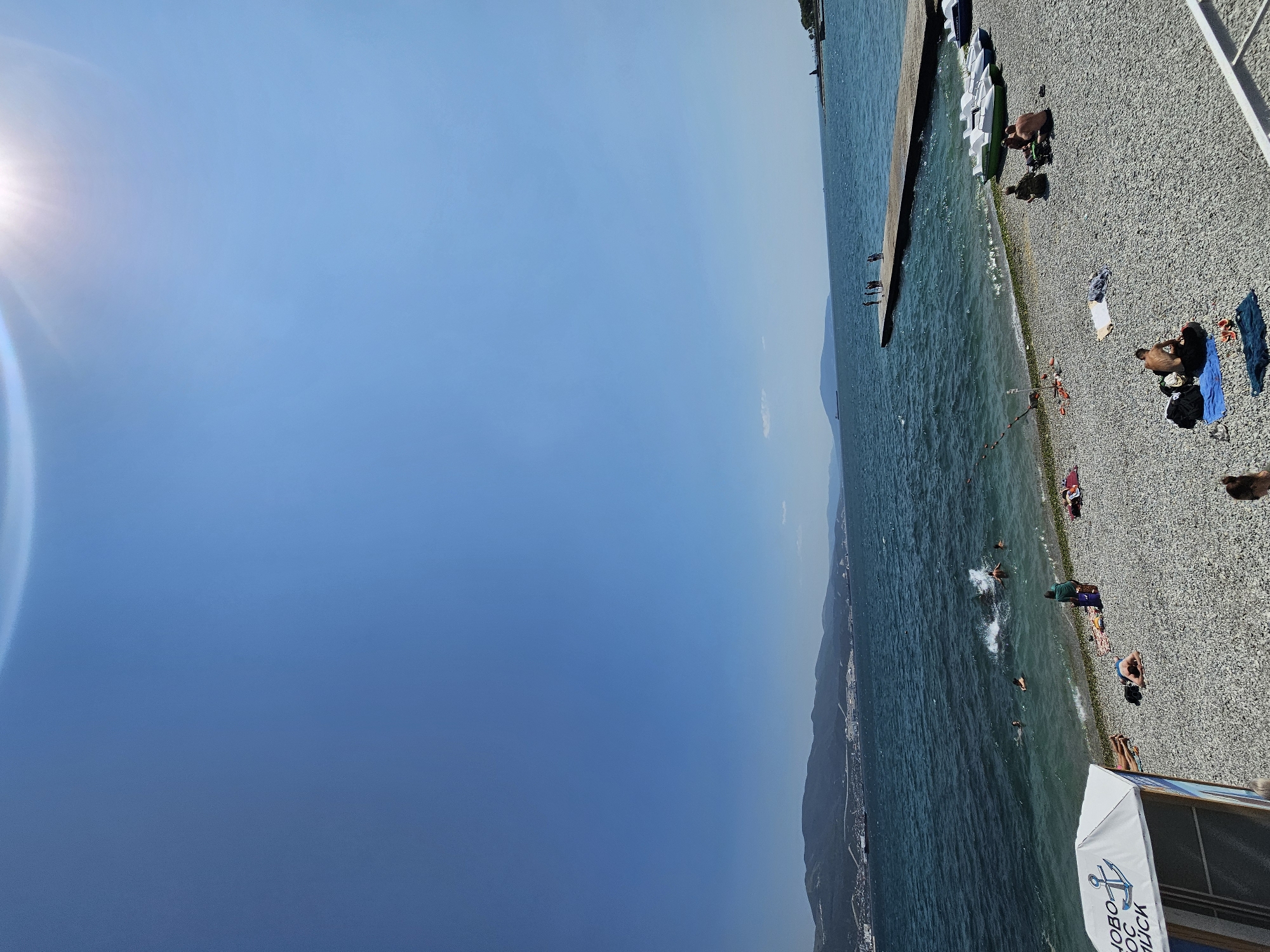 This is a wide-angle, eye-level shot of a pebble beach in Novorossiysk, Russia, on a sunny day. The sea is a vibrant blue, with moderate waves. In the distance, a long pier extends into the water, with several people standing on it. Along the shoreline, a few individuals are swimming and splashing in the shallow water. The beach itself is covered in small, smooth pebbles, with some patches of green algae visible near the water's edge. Several people are relaxing on towels and sunbathing on the beach, while others are sitting in small groups. There are several pedal boats lined up on the sand near the water. In the background, a coastal city with buildings and hills can be seen stretching along the horizon. The sky is a clear, bright blue with a hint of sun flare in the upper right corner. A small, white canopy structure with text and an anchor logo is partially visible in the lower left corner.