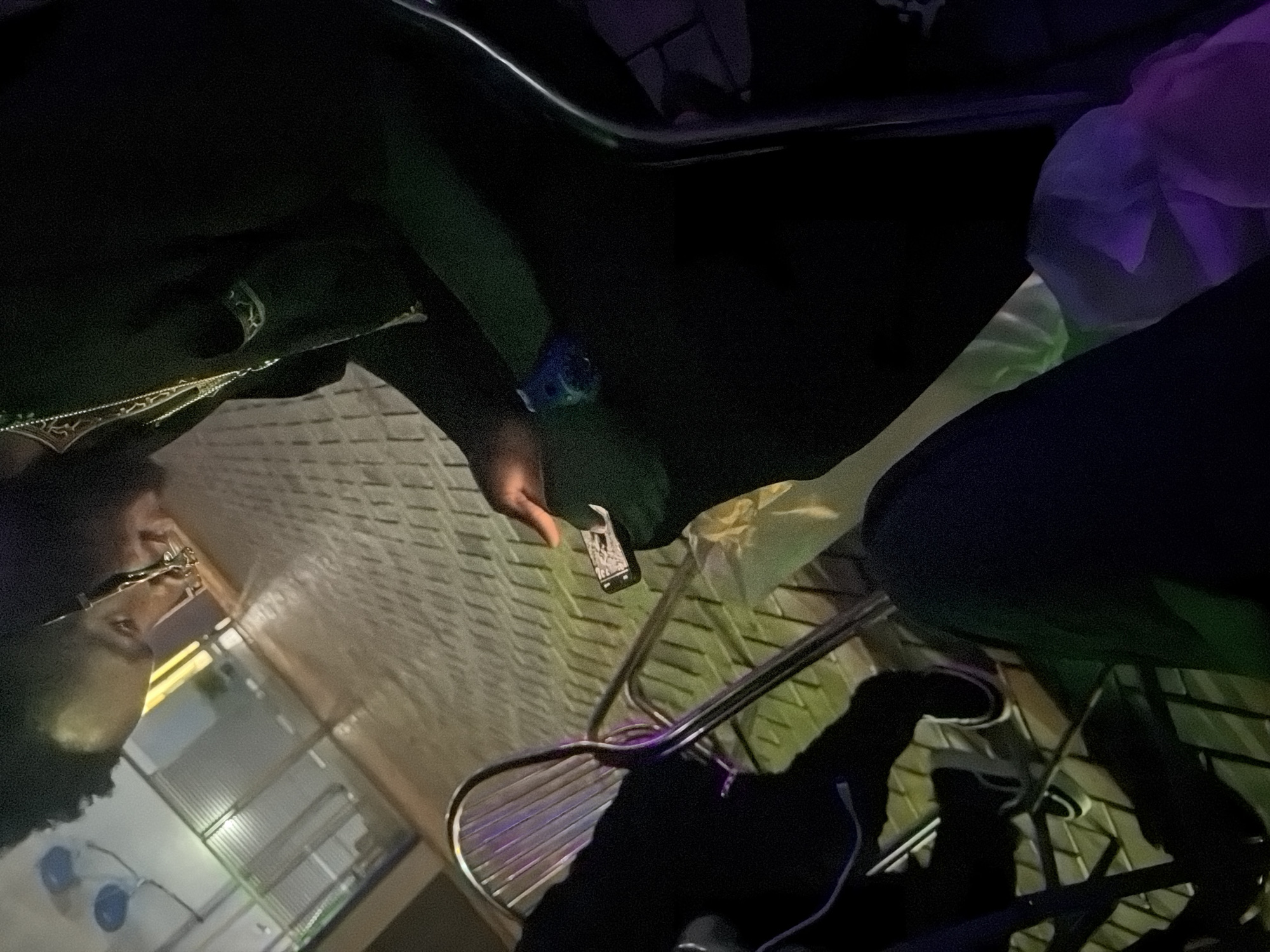 The image shows two individuals in what appears to be an outdoor seating area, possibly a restaurant or a public space. The setting is dimly lit, with warm-toned lighting and some greenish and purplish hues suggesting ambient lighting. The ground is paved with a patterned interlocking surface.
In the foreground, one person is seated, wearing dark clothing and a necklace. They are holding a smartphone and appear to be interacting with it. Their face is partially visible, tilted downwards. Next to them, another person, also in dark clothing, is seated, and their legs are visible. This person is wearing dark pants and white-soled shoes.
Behind the seated individuals, there are metal chairs with a slatted design, and railings. In the background, there are horizontal metal structures that could be part of a fence or a building facade. The overall impression is that of a casual, nighttime gathering. The location in Bukuru, Nigeria, is not explicitly indicated by any visible landmarks or signage within the frame.