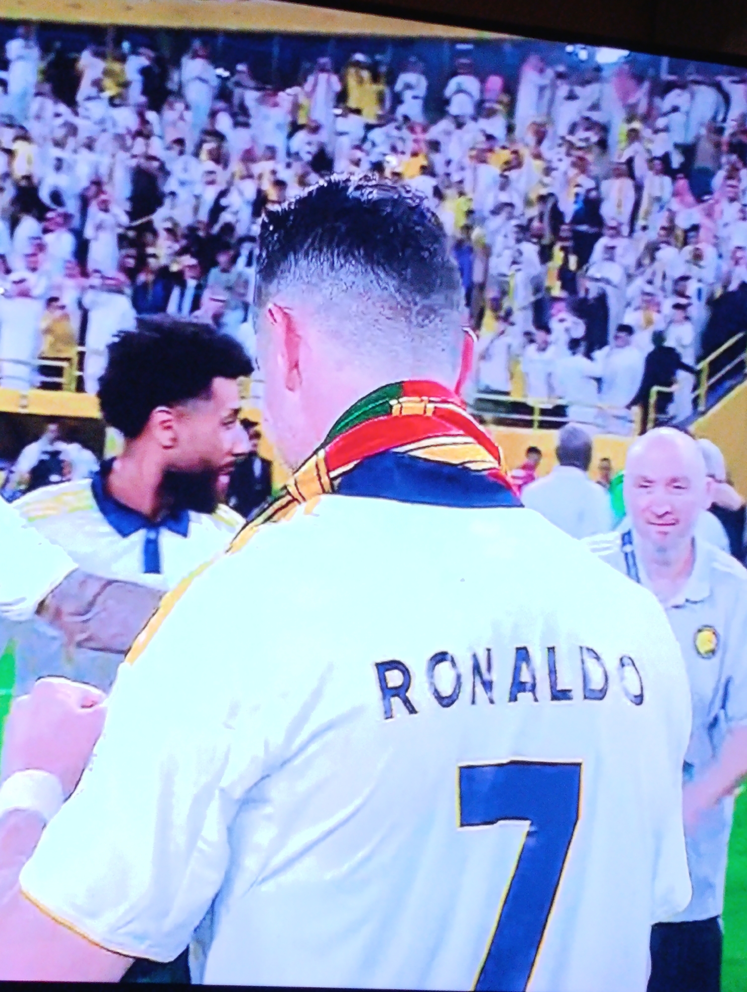 The image shows the back of a person wearing a white jersey with "RONALDO" printed on the back and the number "7" below it. A colorful scarf is draped around the person's neck. To the left is a man with dark curly hair and a beard, wearing a white and blue polo shirt. To the right, another man with a shaved head is wearing a light gray polo shirt with a small yellow logo on the left side. In the background, a large crowd of people is present, many wearing traditional white attire. The overall scene appears to be a celebration or event with a focus on sports. The location is not in the city of Plovdiv, Bulgaria.