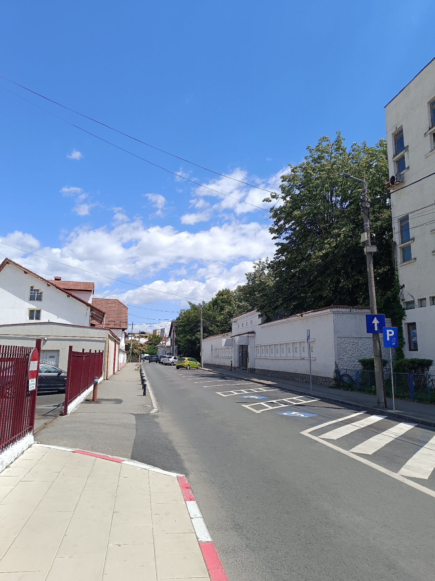 The image depicts a street scene in Târgu Jiu, Romania, under a bright blue sky with scattered white clouds. The perspective is from a sidewalk looking down a paved street lined with buildings on both sides. On the left, a building with white walls and a terracotta tiled roof is visible, featuring windows. A red metal fence runs along the sidewalk in front of this building, with a parked dark-colored car behind it. Further down the street on the left, more buildings with similar architectural styles are seen.
On the right side of the street, a white building with a flat roof and visible air conditioning units is present. A mature tree with lush green foliage is situated behind a white, textured wall in front of this building. Streetlights and utility poles are also visible. Several cars are parked along the right side of the street, including a bright yellow car. The street has a pedestrian crossing marked with white stripes and designated parking spaces marked with blue wheelchair symbols. Traffic signs are also present, including a blue sign with a white arrow pointing upwards and a blue "P" sign indicating parking. The sidewalk is paved with square tiles and has a red and white painted curb. The overall impression is that of a quiet, sunny day in a residential or commercial area of the city.