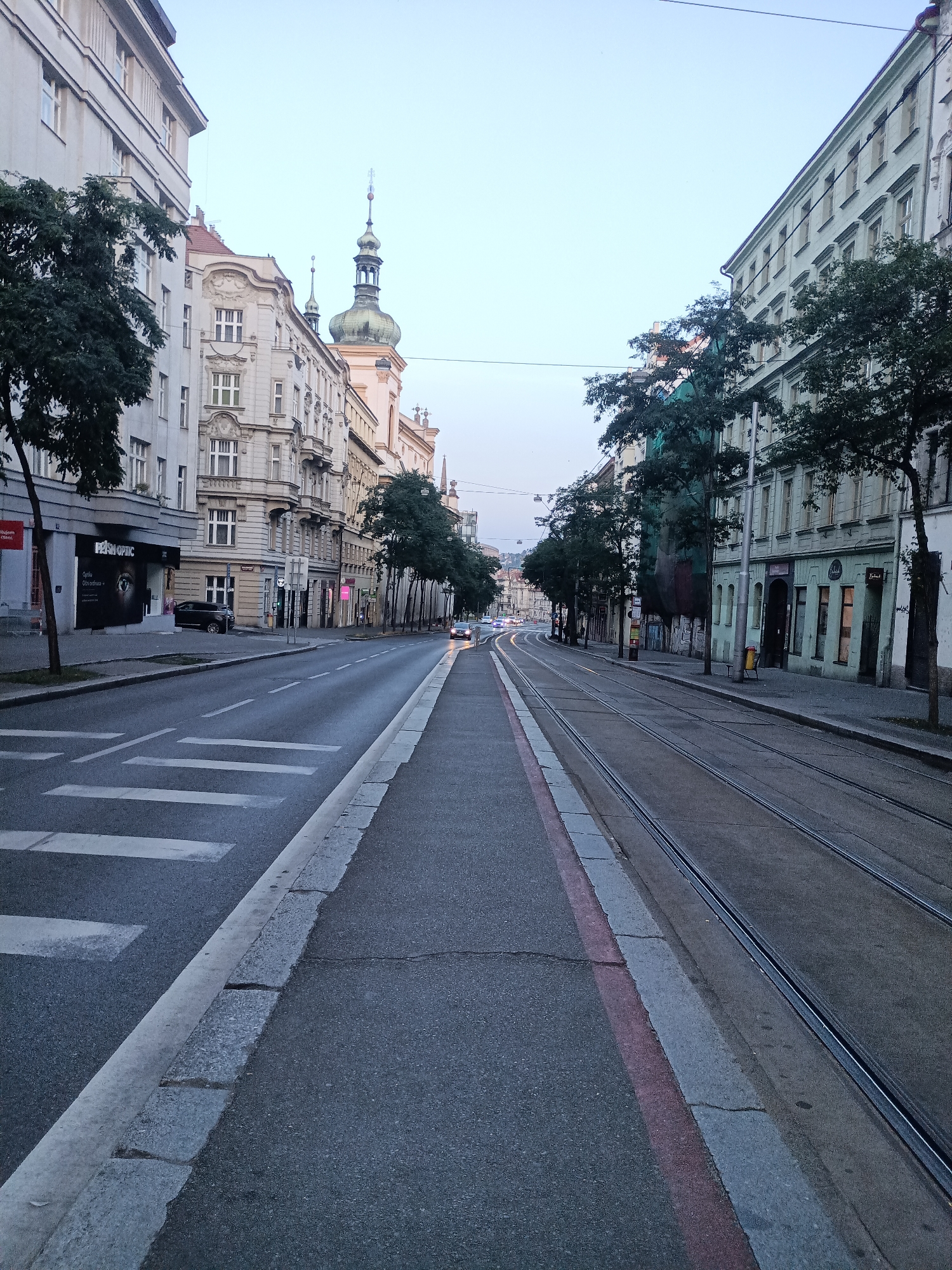 The image shows a street in Prague, Czechia, with tram tracks running down the middle of the road. The street is lined with buildings on both sides, and there are trees along the sidewalks. There is a pedestrian crossing in the foreground. In the background, there are a few cars and a tram. The sky is overcast, and the lighting suggests it is early morning or late evening. The architecture of the buildings appears to be old European style, consistent with Prague. The tram tracks and the general street layout are characteristic of many streets in Prague that are served by public transportation.