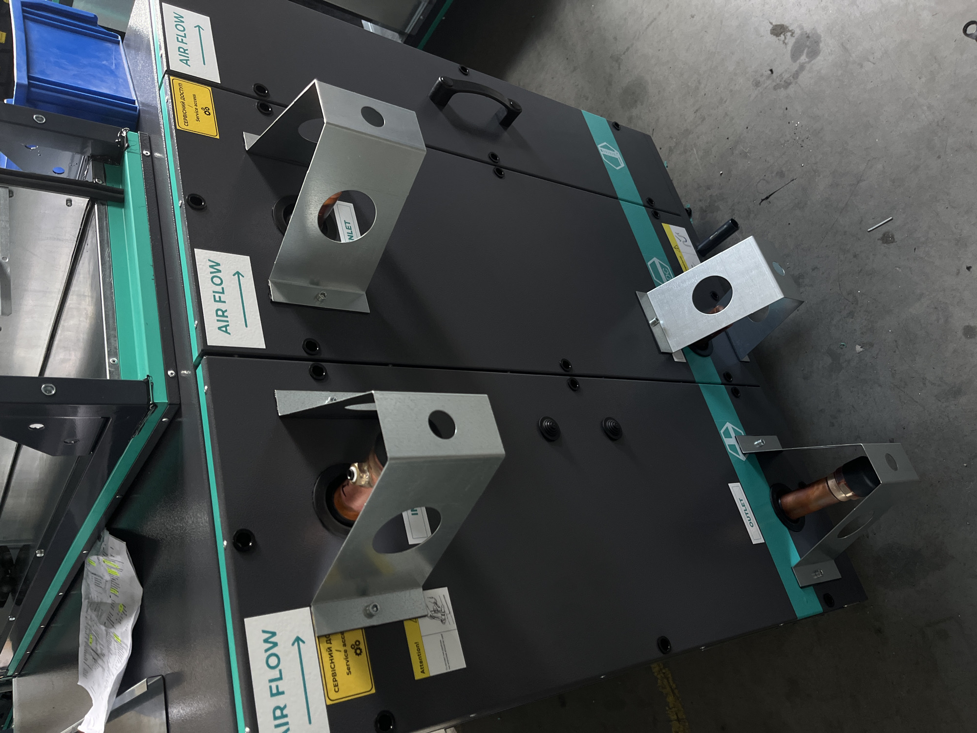 The image shows a close-up of a large, dark gray industrial machine with teal accents. On the left side of the machine, a white sticker with an arrow pointing right reads "AIR FLOW". Below this, a yellow sticker has Ukrainian text that translates to "Service access" and includes an icon of gears. Further down, another white sticker reads "Attention!".
To the right of the "AIR FLOW" sticker, there is a circular opening labeled "INLET" with a copper pipe fitting visible inside. Above this opening is a metallic bracket.
Further to the right, another "AIR FLOW" sticker with a right-pointing arrow is visible. A black handle is mounted on the side of the machine.
At the bottom of the machine, there is another opening labeled "OUTLET" with a copper pipe fitting extending out, supported by a metallic bracket.
The machine appears to be situated in a warehouse or industrial setting, with a concrete floor visible. The background is slightly out of focus, but shows parts of the machine and possibly other industrial equipment. There are no people visible in the image. The overall impression is of a functional piece of industrial equipment. There is no information in the image to suggest a specific location within Kyiv, Ukraine.