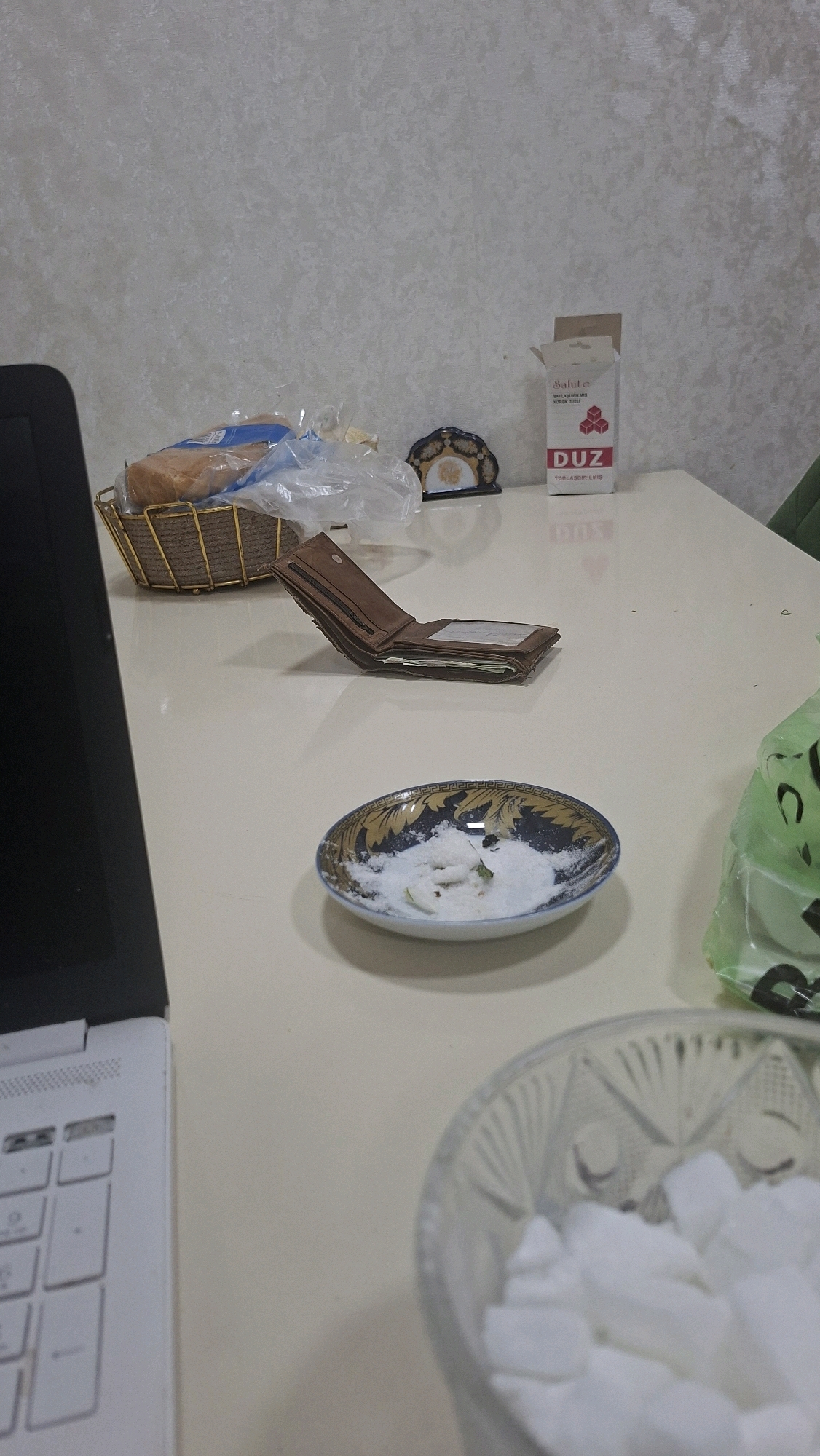 The image depicts a table with various objects. On the left, a white laptop is partially visible. In the center, a brown leather wallet is open, revealing some contents. Next to the wallet, a small decorative dish contains white powder and some green items. Behind the wallet, a woven basket holds bread wrapped in plastic. To the right of the basket, a small cardboard box with text and an image of cubes is present. Further to the right, a green plastic bag with a cartoonish face is visible. The scene appears to be indoors, with a textured wall in the background. The presence of everyday objects suggests a domestic or office setting. There is no indication of the city of Mingachevir, Azerbaijan, in the provided image.