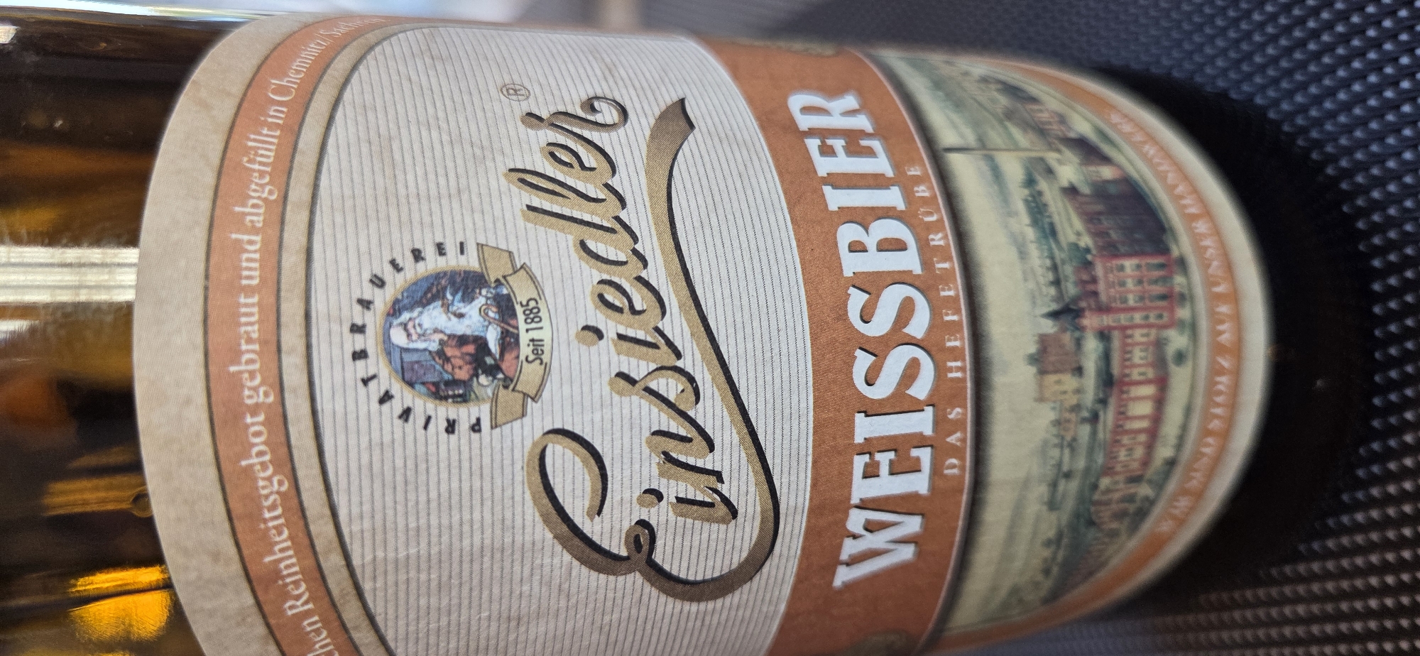 A close-up image captures the label of an "Einsiedler Weissbier" beer bottle. The label is light beige with horizontal lines, framed by a rust-orange band. At the top, curved text reads, "...chen Reinheitsgebot gebraut und abgefüllt in Chemnitz/Sachsen," indicating the beer is brewed and bottled in Chemnitz, Saxony, Germany. Below this, a circular emblem shows a bearded man, possibly a hermit, holding a staff, with "PRIVATBRAUEREI" (Private Brewery) curving above and a banner reading "Seit 1885" (Since 1885) beneath. The brand name "Einsiedler" is prominently displayed in a gold, stylized script, underscored by a gold ribbon. Below this, "WEISSBIER" (White Beer) is written in large white capital letters, with "DAS HEFETRÜBE" (The Yeast Cloudy) in smaller text directly underneath. The bottom section of the label features a detailed, panoramic illustration of a historical townscape or brewery complex with red-roofed buildings and trees. Partially visible text along the bottom edge states "...SIND STOLZ AUF UNSER HANDWERK" (are proud of our craftsmanship).