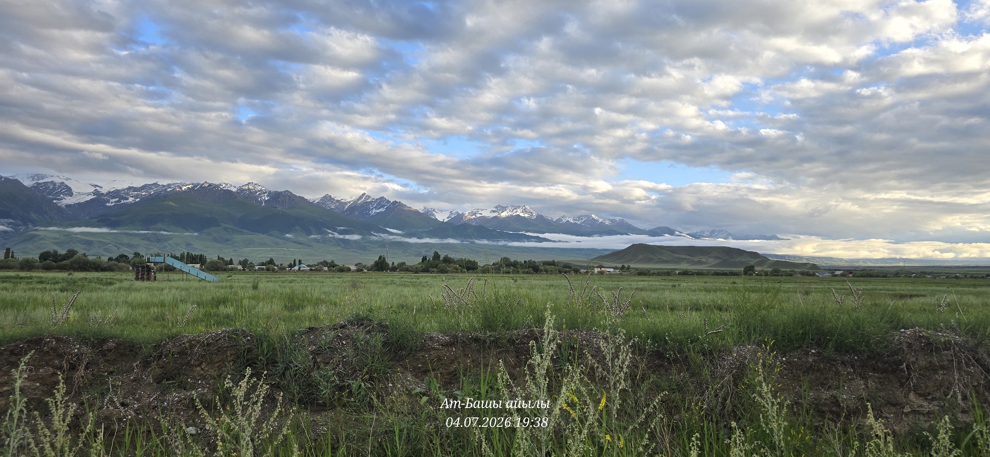 The image shows a wide landscape in At-Bashy, Kyrgyzstan. In the foreground, there is a grassy field with some dry brush and soil banks. A small, blue playground slide structure with wooden supports is visible in the middle ground, situated within the grassy field. Beyond the field, a line of trees separates it from a distant village with scattered buildings. In the background, a range of majestic, snow-capped mountains dominates the horizon, partially obscured by low-lying clouds. The sky is filled with dramatic, textured clouds, with patches of blue sky visible. The overall scene is a natural landscape with signs of human presence in the form of the playground and village. Text overlays indicate "Ат-Башы айылы" (At-Bashy village) and a date and time: "04.07.2026 19:38".