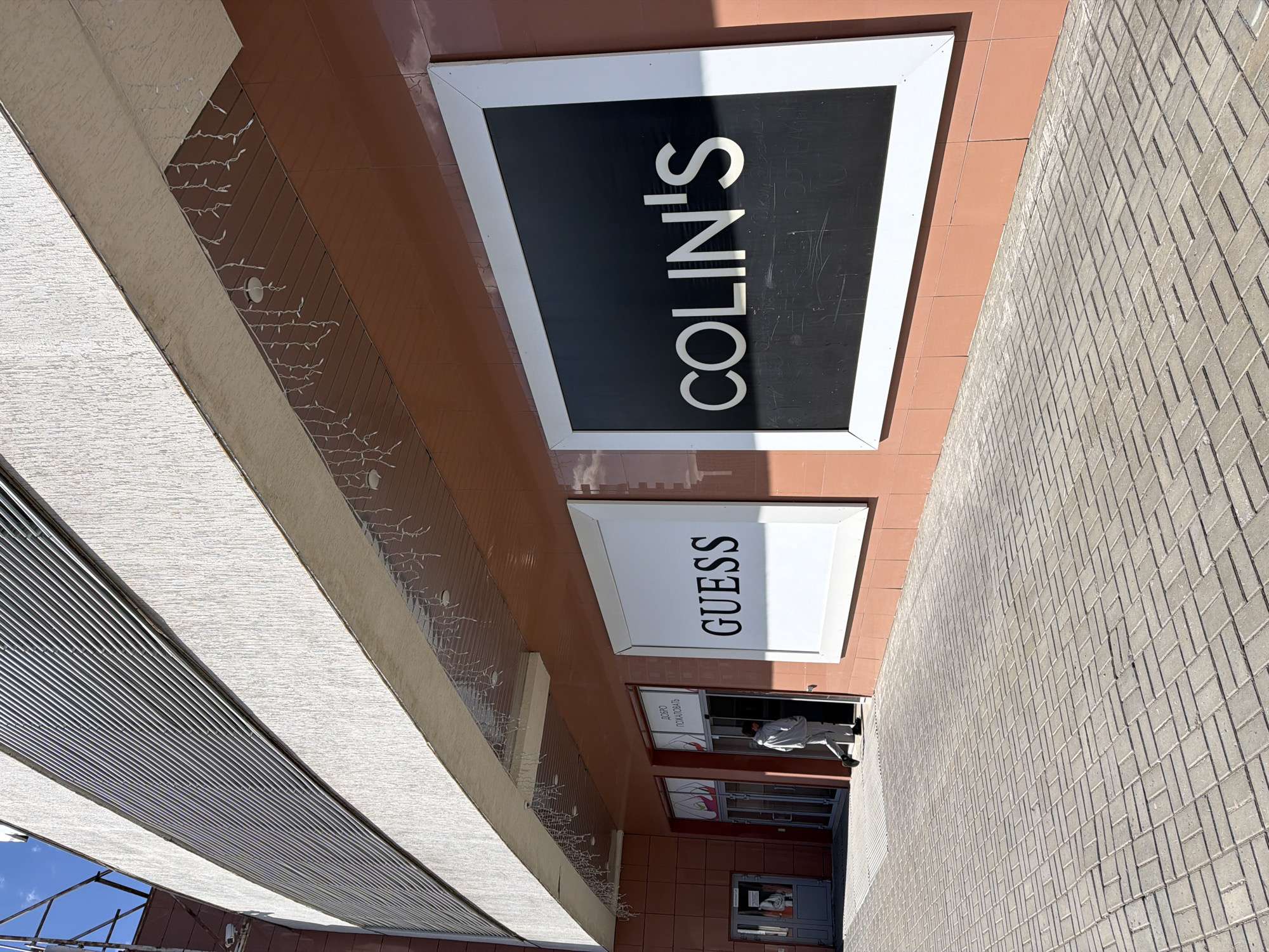 The image shows the exterior of a building in Izhevsk, Russia, with signs for "COLIN'S" and "GUESS" visible. A person is seen near the entrance to the store. The building has a brown facade with decorative lights and a sidewalk made of gray bricks. An awning structure is visible overhead, with a glimpse of a blue sky in the background.