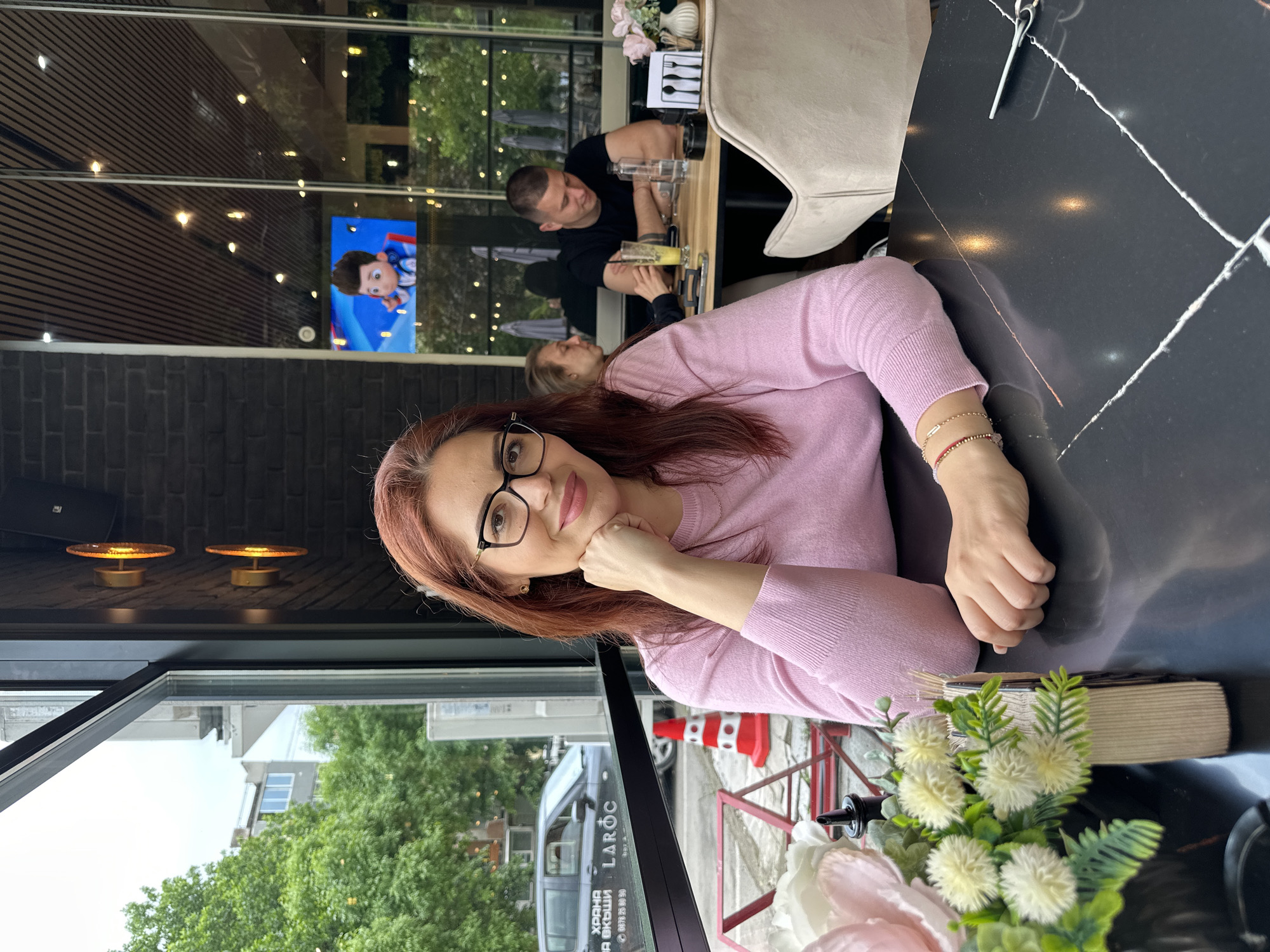 The image captures a woman seated at a black marble-patterned table in a restaurant or cafe in Montana, Bulgaria, as suggested by the "LAROC" van with Cyrillic text visible through the window. The woman, with reddish-brown hair and glasses, wears a pink sweater and rests her chin on her hand. On the table, there is a floral arrangement and a stack of napkins. In the background, other patrons are present; a man wearing a black shirt is seated at a table with a woman. A television screen displays a cartoon character, reflecting in the glass of the window. The restaurant has a modern interior with black brick walls and decorative wall lighting. Red and white traffic cones and greenery are visible outside through the window, indicating an urban setting.