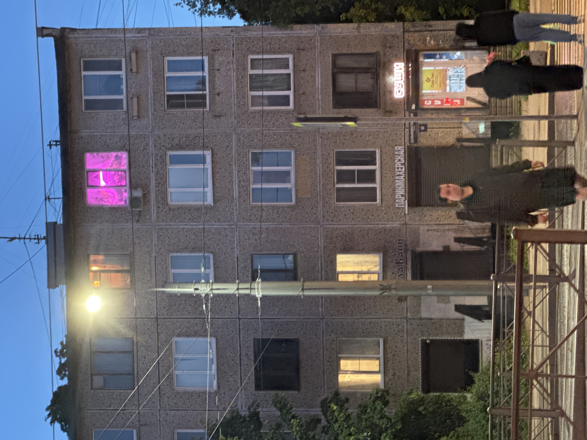 An exterior, medium shot captures a multi-story, grey, concrete apartment building at dusk in Saint Petersburg, Russia. The building has numerous windows, some illuminated with warm light, while one window on the third floor from the top emanates a vibrant pink glow. A street light casts a bright yellow light on the scene. Below the windows on the ground floor, two signs are visible: one reads "ПАРИКМАХЕРСКАЯ" (Hairdresser) and the other, a neon sign, says "СУШИ" (Sushi). Several people are walking on the sidewalk in front of the building, some crossing a street at a pedestrian crossing. The sky is a deep blue, indicating the transition from day to night.