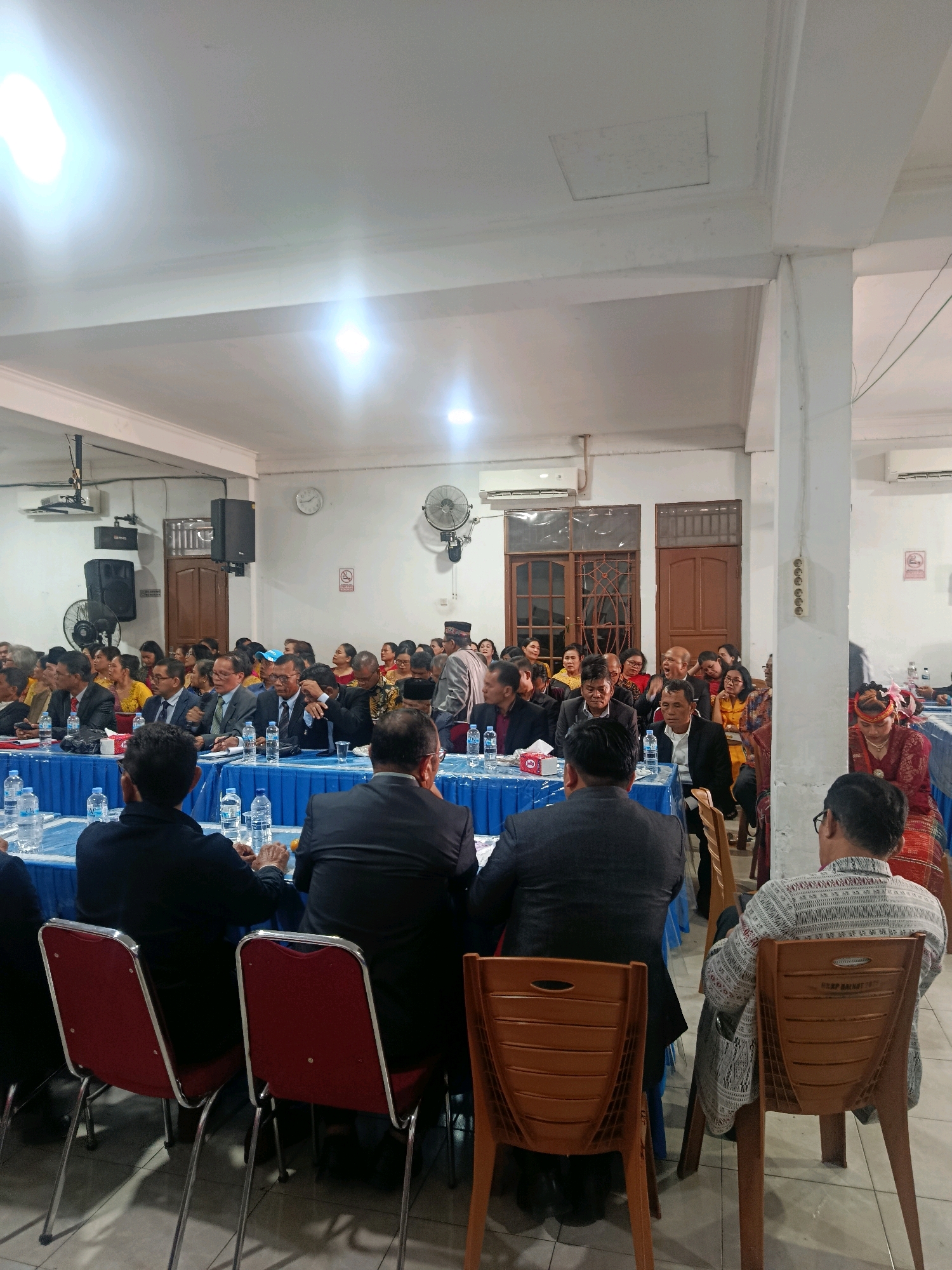 The image depicts an indoor gathering in Babelan, Indonesia. A group of people are seated at tables covered with blue tablecloths. Some individuals are wearing formal attire, including suits and ties, while others are in more traditional clothing. There are bottles of water and tissues on the tables. In the background, there are doors, windows, and what appear to be air conditioning units mounted on the wall. A fan is also visible. The lighting suggests the event is taking place in the evening or indoors with artificial illumination. The atmosphere seems to be one of a formal meeting or celebration.