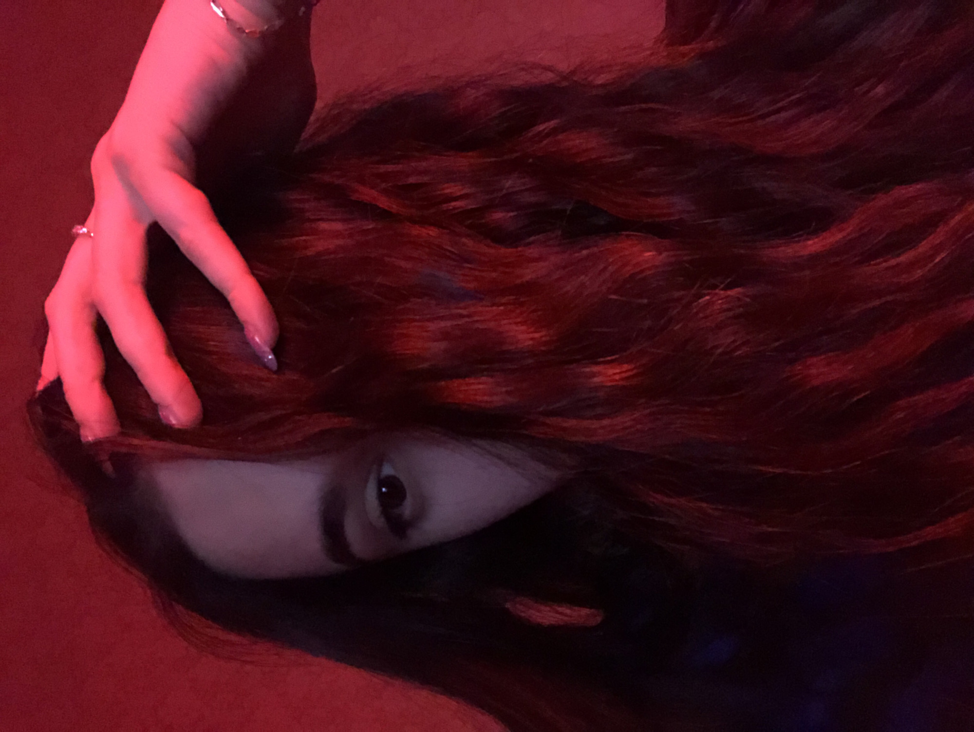 A close-up, horizontally oriented image features a person with long, dark red-brown, wavy hair. A hand with slender fingers and pale skin rests on the hair, near the left side of the frame. The person's face is partially obscured by the hair, but one eye is visible, looking towards the viewer. The lighting in the scene is predominantly red, giving the image a somewhat dark and mysterious appearance. No information is available to suggest that the image was taken in Noviy, Russia.