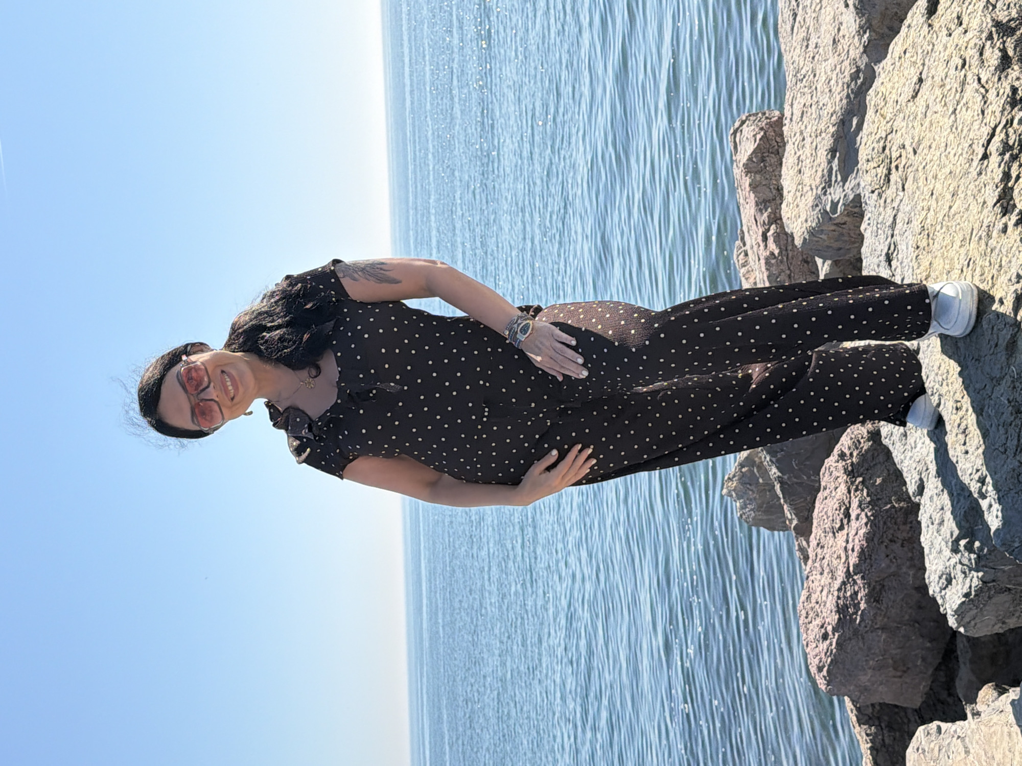 The image shows a woman standing on rocks overlooking the sea. The woman is wearing a black jumpsuit with white polka dots and white shoes. She has dark hair and is wearing sunglasses. She has tattoos on her left arm. Her hands are resting on her belly. The rocks she is standing on are grey and appear natural. The sea is calm, and the sky is clear. The location is likely in Kadıköy, Turkey, given the presence of the sea and rocks that are typical of the area's coastline.