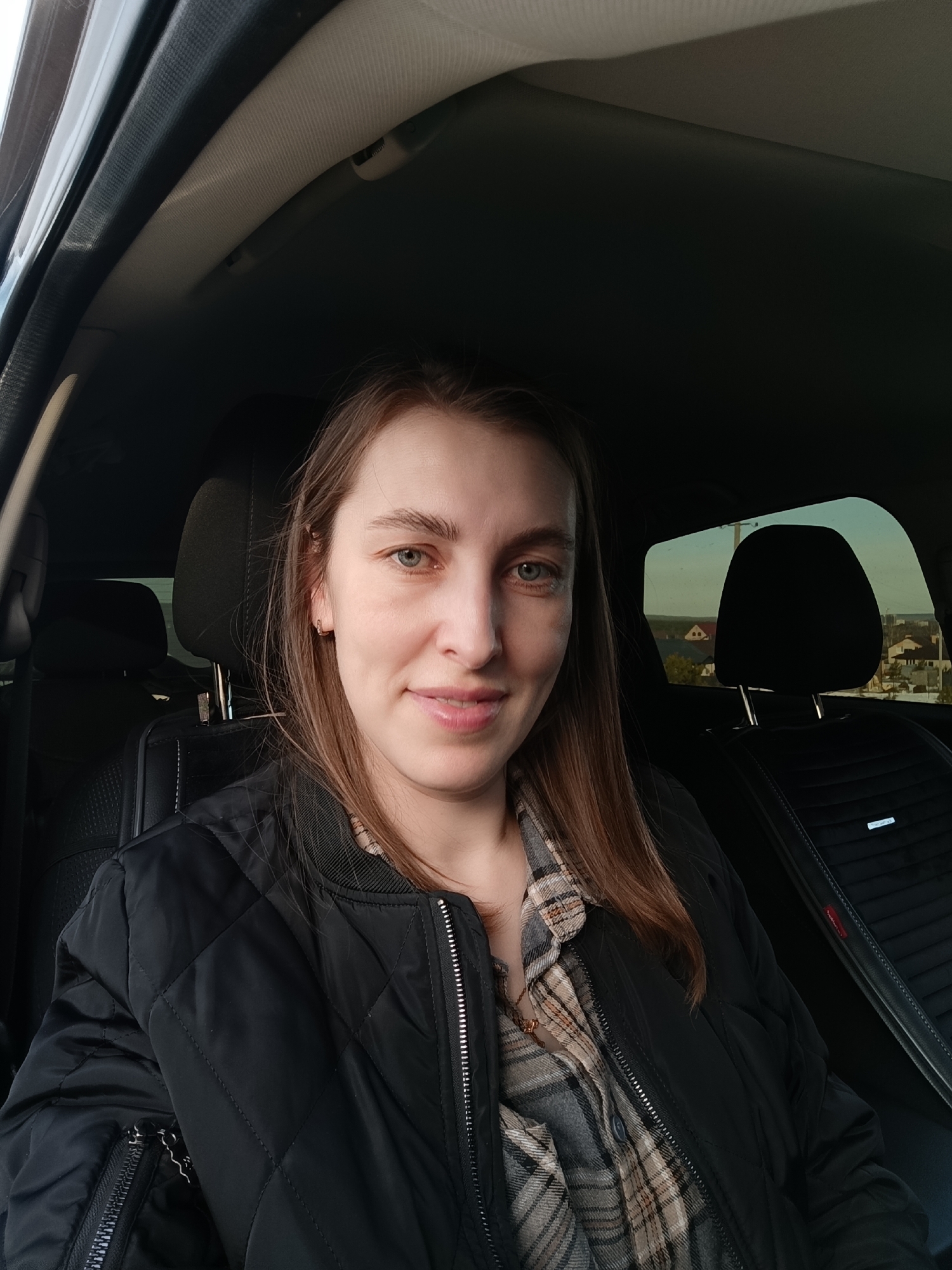 A young woman is seated in the driver's seat of a car. She is wearing a black quilted bomber jacket over a plaid flannel shirt. Her hair is light brown and shoulder-length. The car's interior is black leather. Outside the passenger-side window, a residential area is visible, suggesting a location in the city of Chaikovsky, Russia. The view includes houses and trees against a slightly overcast sky.