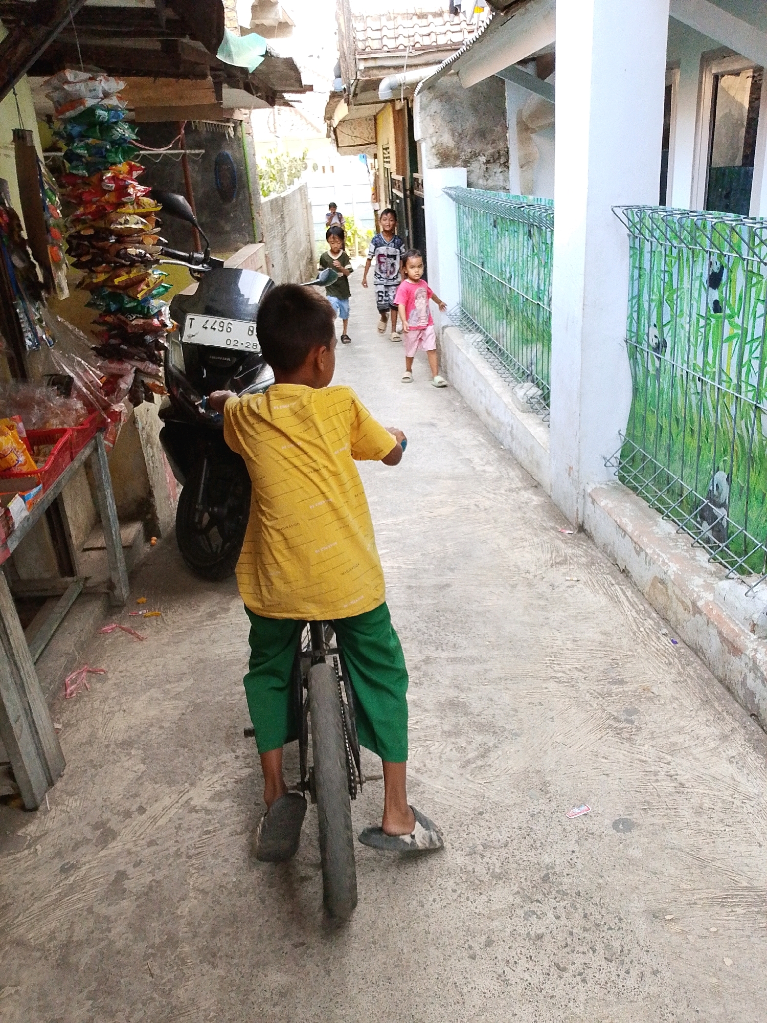 In Karawang, Indonesia, a young boy wearing a yellow shirt with a patterned design and green pants rides a bicycle down a narrow, paved alleyway. He is positioned in the foreground, facing away from the viewer and towards other children further down the alley. Behind him, a black motorcycle is parked.
Several other children are visible in the mid-ground. Two boys, one in a green shirt and blue shorts and another in a white and black patterned shirt, are walking in the same direction as the boy on the bicycle. A young girl in a pink shirt and shorts is also walking in the alley.
The alley is lined with buildings and structures. On the left side, there is a stall with various items hanging, including colorful bags of snacks, and a red plastic bin. On the right side, a white wall with a green metal fence separates the alley from a residential area. The fence is decorated with images of pandas among bamboo. The ground is made of textured concrete. The overall scene depicts a typical street scene in a residential area with children playing and engaging in everyday activities.