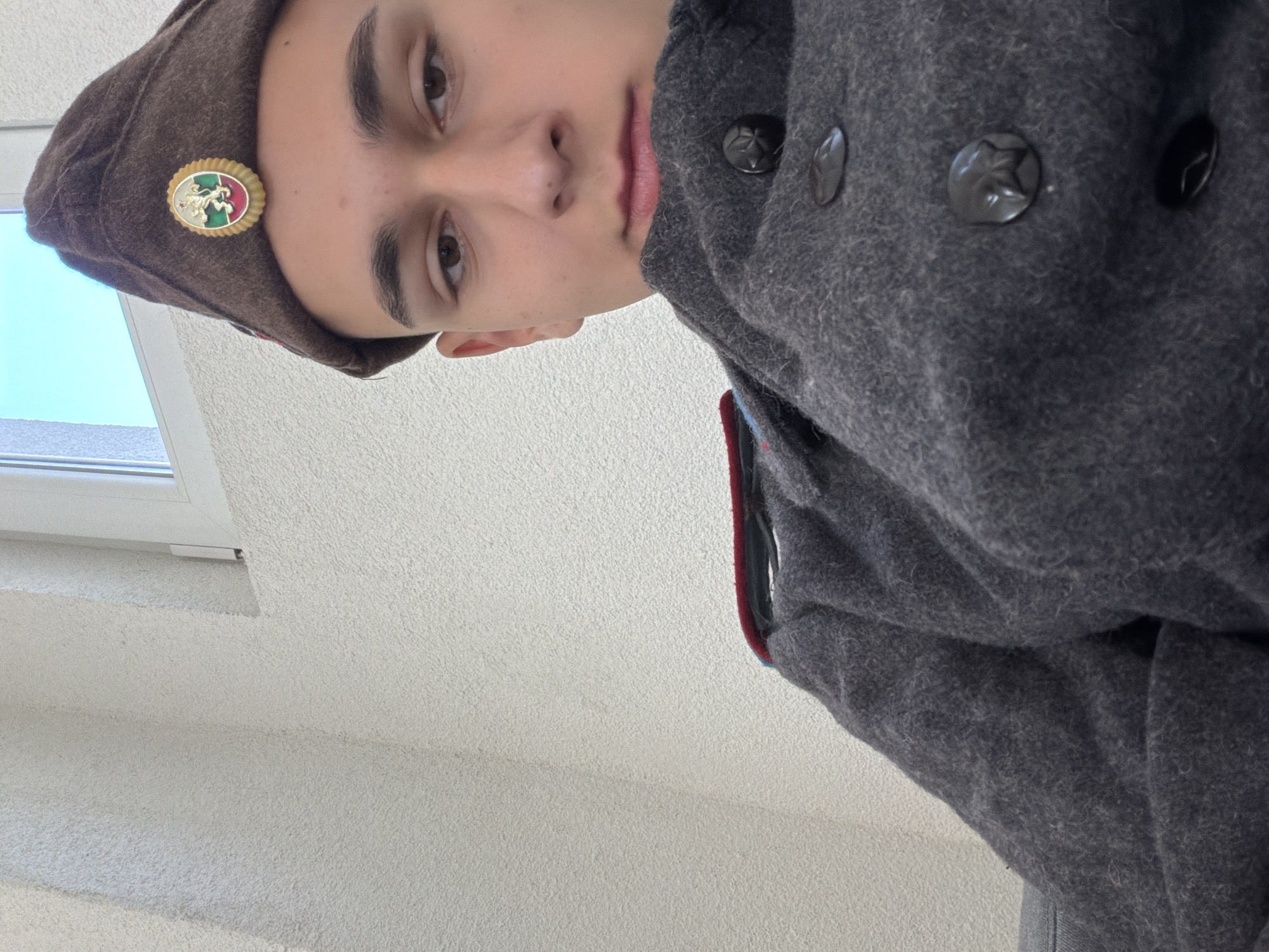 A young person is depicted in a close-up, angled shot. The individual is wearing a brown beret adorned with a circular badge featuring a heraldic design, and a gray wool coat with buttons marked with a star. The photo was taken in Shumen, Bulgaria. The foreground is filled with the textured wall and coat. A window is visible in the background.