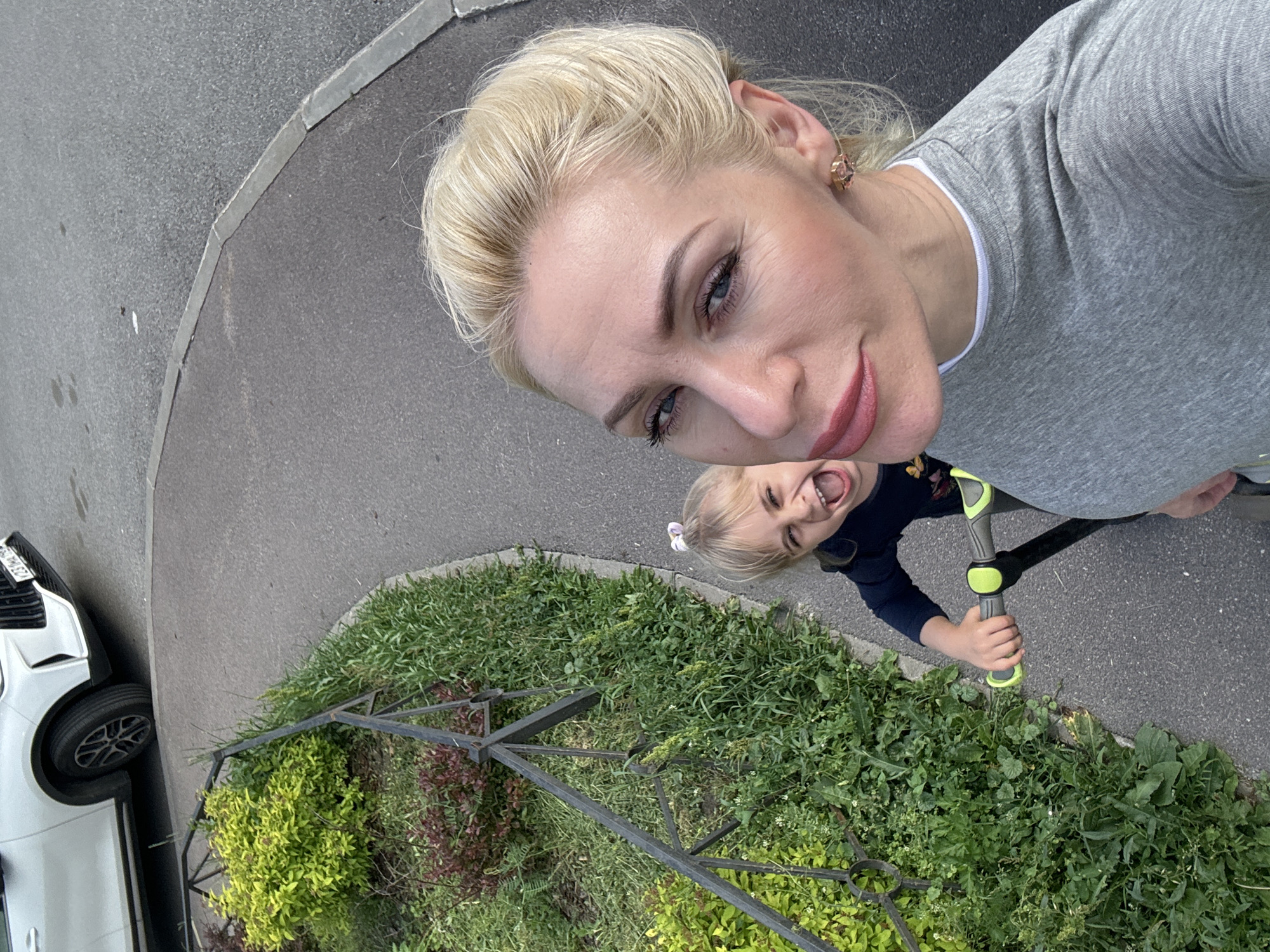 In a top-down selfie taken outdoors, a blonde woman in a grey t-shirt captures her face in the foreground. Her hair is pulled back, and she wears subtle makeup with pink lipstick and mascara. To her left and slightly behind her, a young blonde child with a hair clip rides a scooter. The child has an open-mouthed expression of excitement. They are positioned on a paved pathway bordered by a green grassy area and a black metal fence. In the background, the front of a white SUV is visible, parked on asphalt. The scene suggests a casual outdoor activity in an urban or suburban environment.