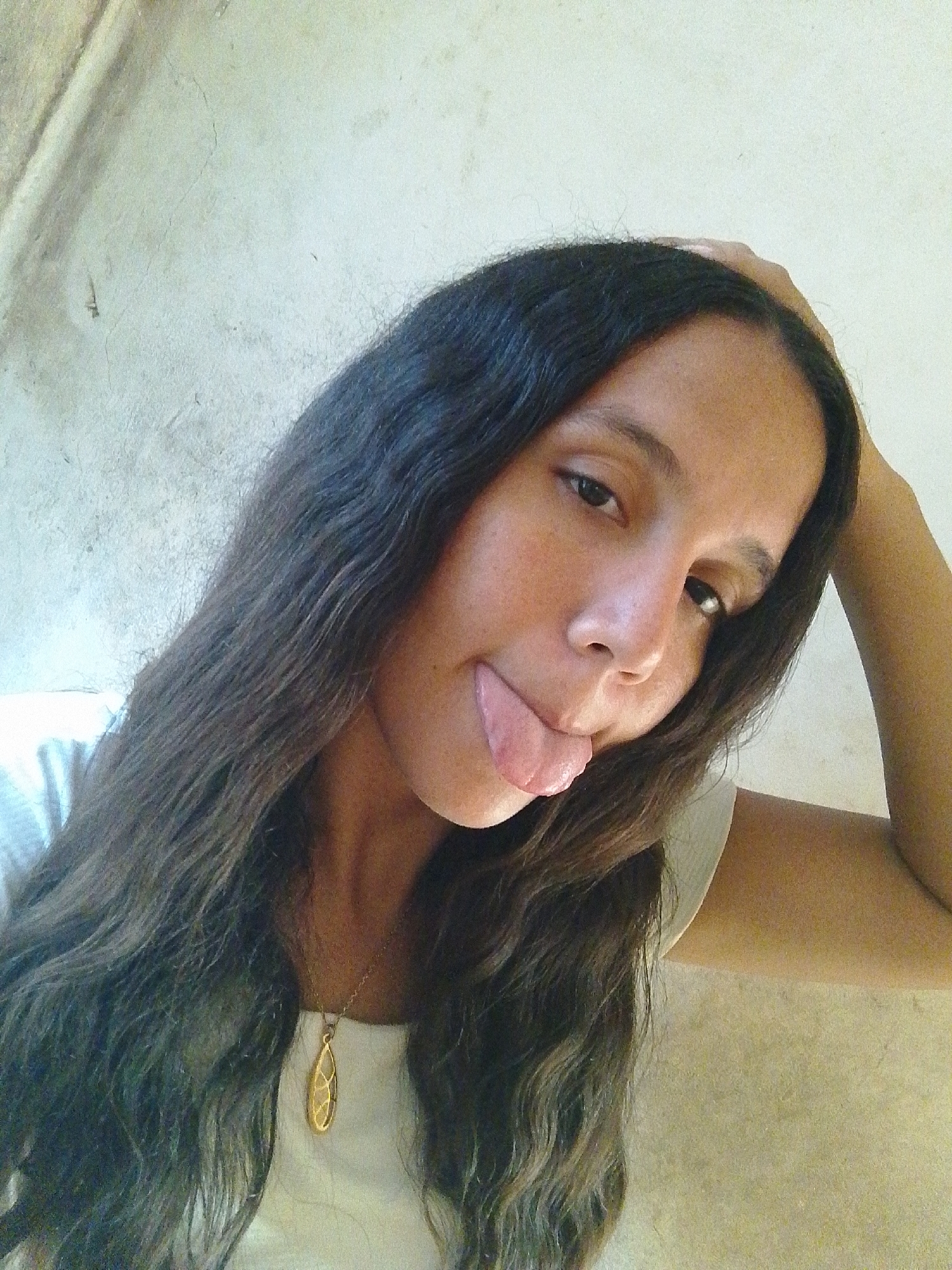 A woman with long dark hair, a gold pendant necklace, and a white top sticks her tongue out at the camera. She has one hand on her head. The background appears to be a plain, light-colored wall. There is no immediately identifiable information to pinpoint the location as Juan Griego, Venezuela.