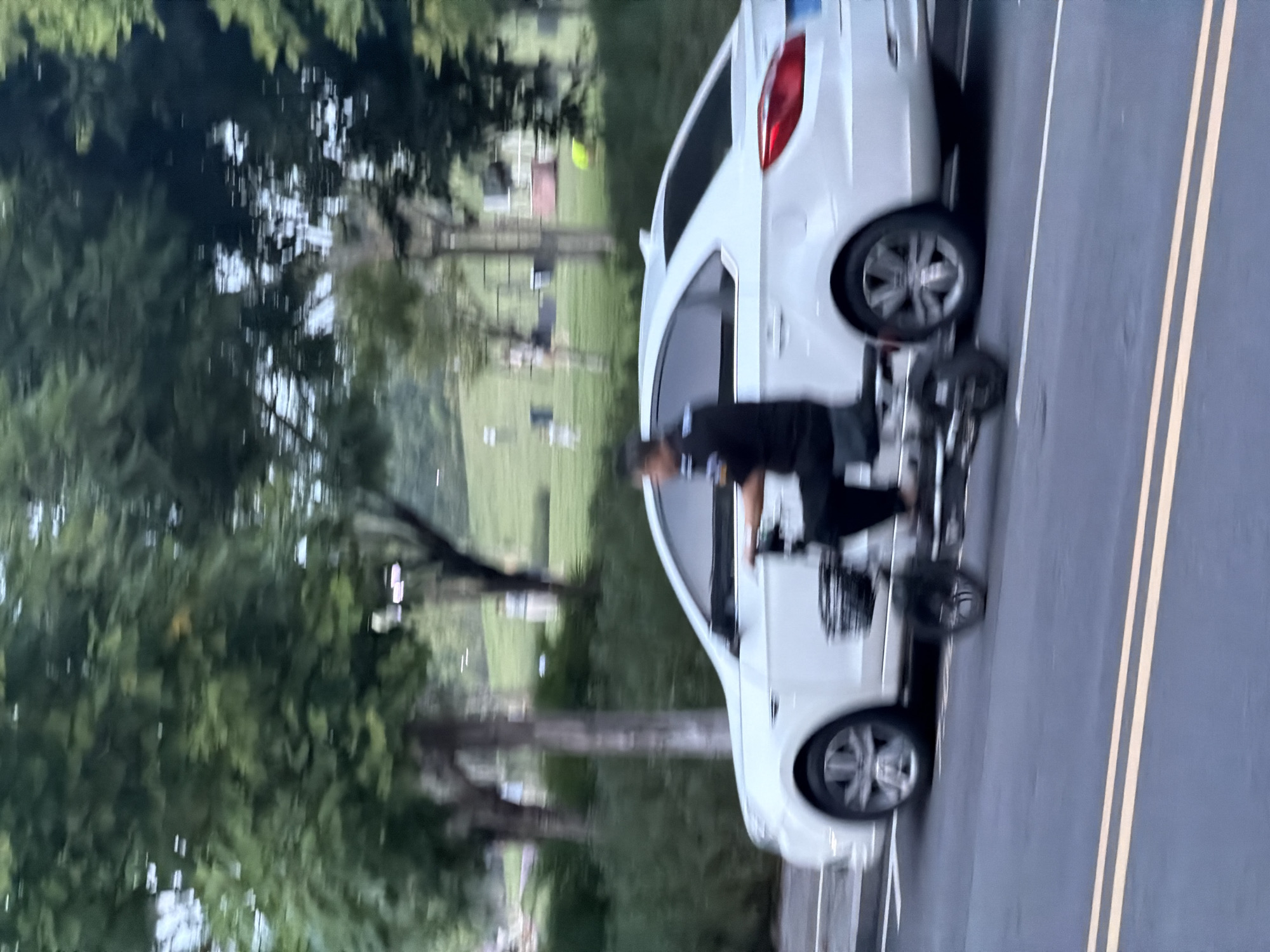 A motion-blurred, medium shot captures a person riding a three-wheeled electric scooter alongside a white sedan on a paved road in the city of Jiangjin, China. The scooter rider, wearing a dark shirt and pants, is positioned on the left side of the frame. The white sedan is on the right, with its rear end closer to the viewer. The background features trees and a grassy slope with some faint structures. The road has yellow lane markings. The overall scene is slightly hazy, suggesting a possible overcast day or low light conditions.