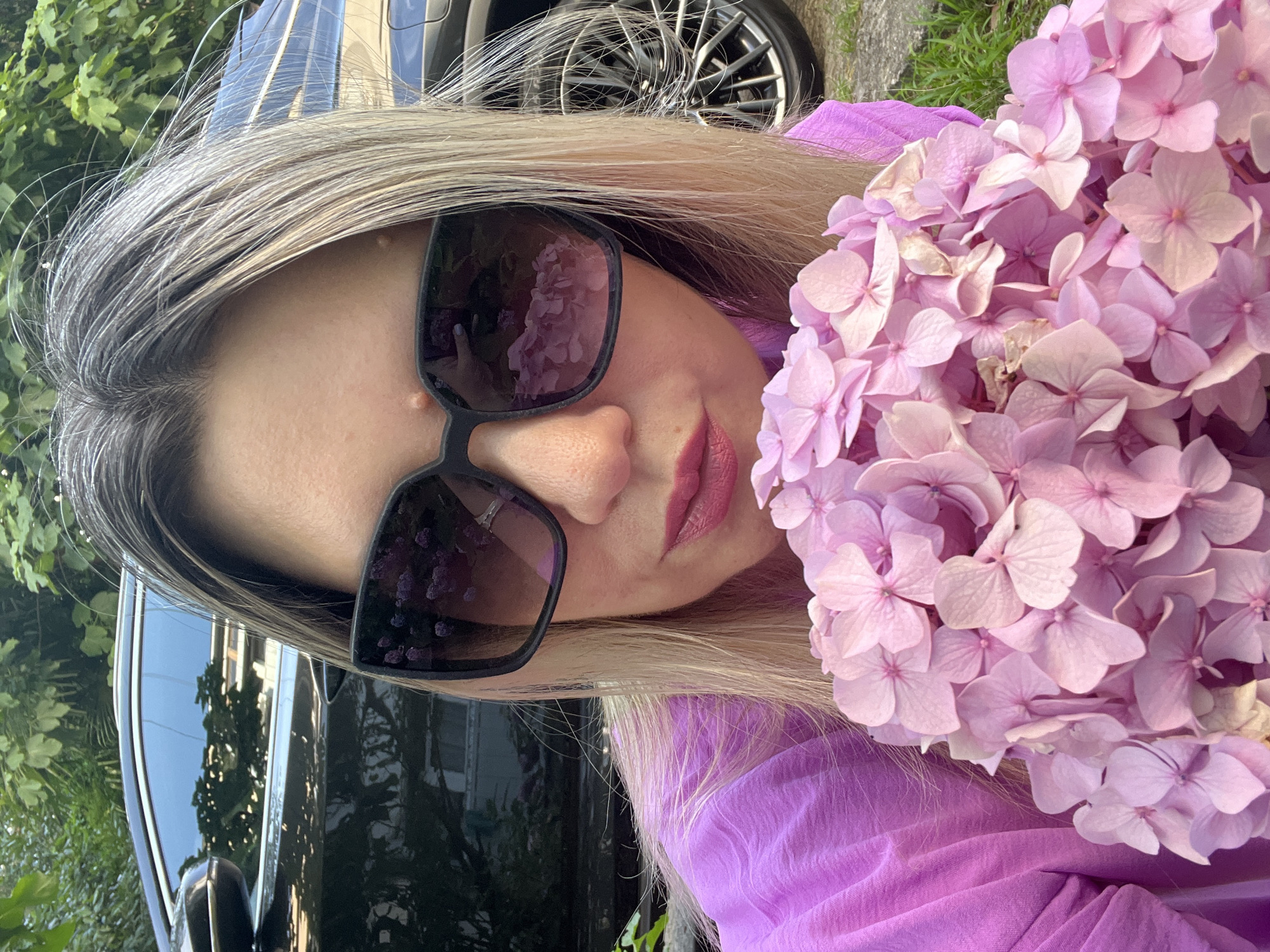 A woman with blonde hair, wearing large dark sunglasses and a purple top, holds a bouquet of pale pink hydrangeas. The flowers are the focus of the foreground, with their delicate petals creating a soft texture. The woman's face is partially visible, with her lips painted a muted berry color. The sunglasses reflect an image of the flowers, creating a mirrored effect. In the background, a portion of a dark car is visible, with its shiny surface reflecting the surrounding greenery and a building. The scene appears to be outdoors, possibly in a garden or residential area, given the presence of plants and a car. The lighting suggests it is daytime, with natural light illuminating the scene. The overall mood is relaxed and pleasant. There is no visible text in the image.