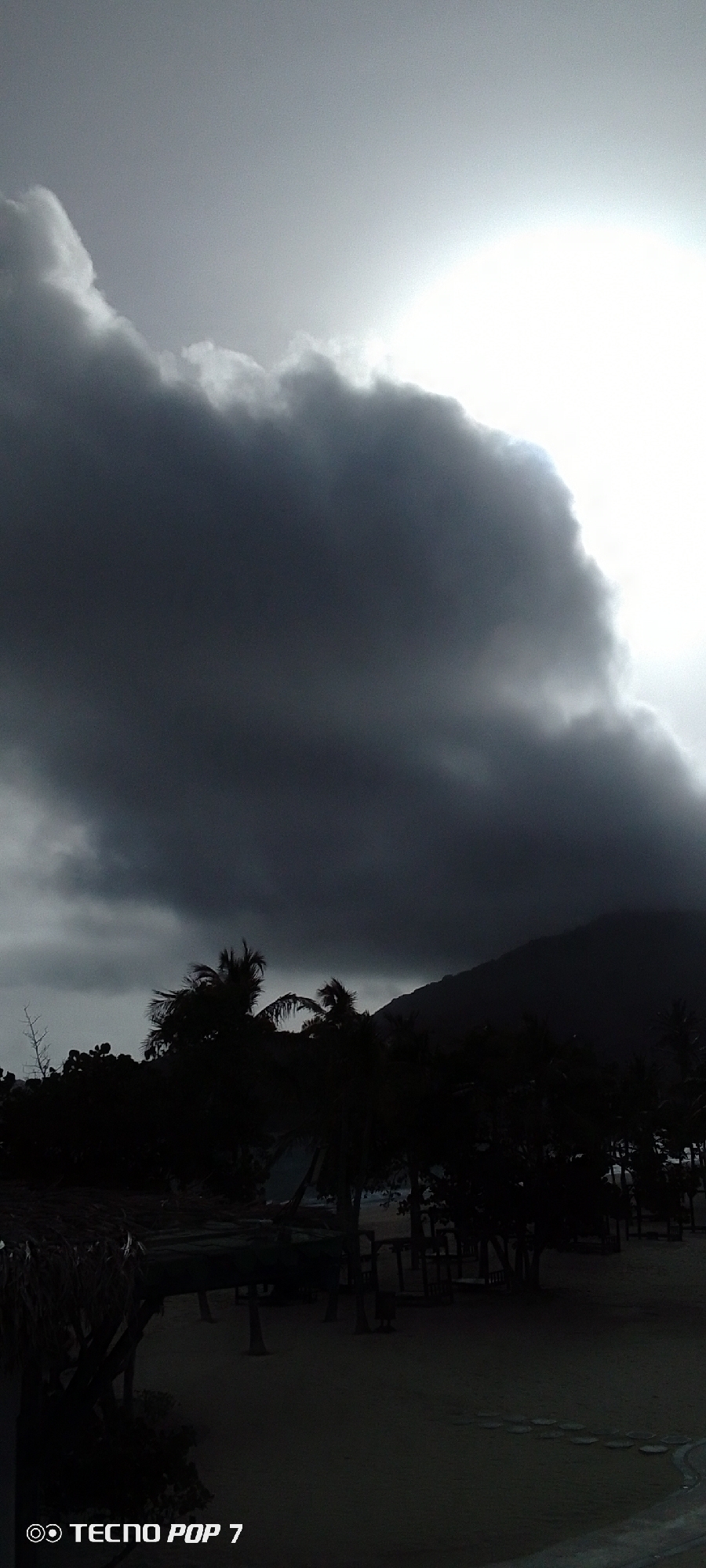 The image, taken in Pedro González, Venezuela, depicts a dark, overcast scene at what appears to be a beach. Heavy, dark clouds dominate the sky, obscuring the sun. Silhouetted trees, possibly palm trees, are visible in the foreground. A dark mountain or hill is visible in the middle ground, further back from the vegetation. The foreground also contains structures that could be benches or shaded areas on the beach. The scene is generally dark with minimal detail due to the low lighting and weather conditions. The camera model, "TECNO POP 7," is printed in the bottom left corner of the image.