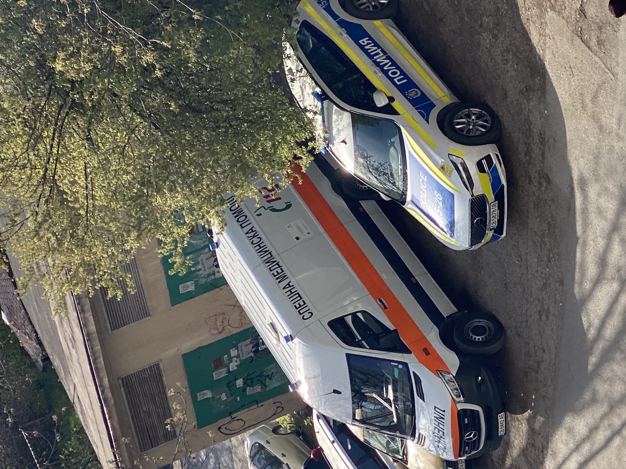 This media file captures an outdoor scene in Pleven, Bulgaria, showing two emergency vehicles parked on a paved surface under bright, direct sunlight, indicating daytime. The image is taken at an oblique angle, with the vehicles appearing rotated counter-clockwise.
In the center, a white ambulance with an orange stripe running horizontally along its side is prominently displayed. The words "СПЕШНА МЕДИЦИНСКА ПОМОЩ" (Emergency Medical Aid) are visible in Cyrillic on its side, and "112" is partially seen. The front license plate of the ambulance reads "CB 8442 AE".
Behind the ambulance, a white police car with blue and yellow markings is parked. The word "ПОЛИЦИЯ" (Police) in Cyrillic is visible on its side, along with "POLICE" in English and the numbers "15-26". Its front license plate displays "CB 5878 OB".
To the left of the vehicles, a building with a beige facade and green painted sections is visible. There are several rectangular, possibly boarded-up or shuttered, windows. Graffiti and various small notices or posters are stuck to the building's walls. Lush green foliage from a tree, likely in full leaf, extends over the upper left portion of the scene, with sunlight filtering through its leaves. The ground is a light-colored paved area, and shadows are sharp, indicating clear weather. The vehicles appear to be stationary.