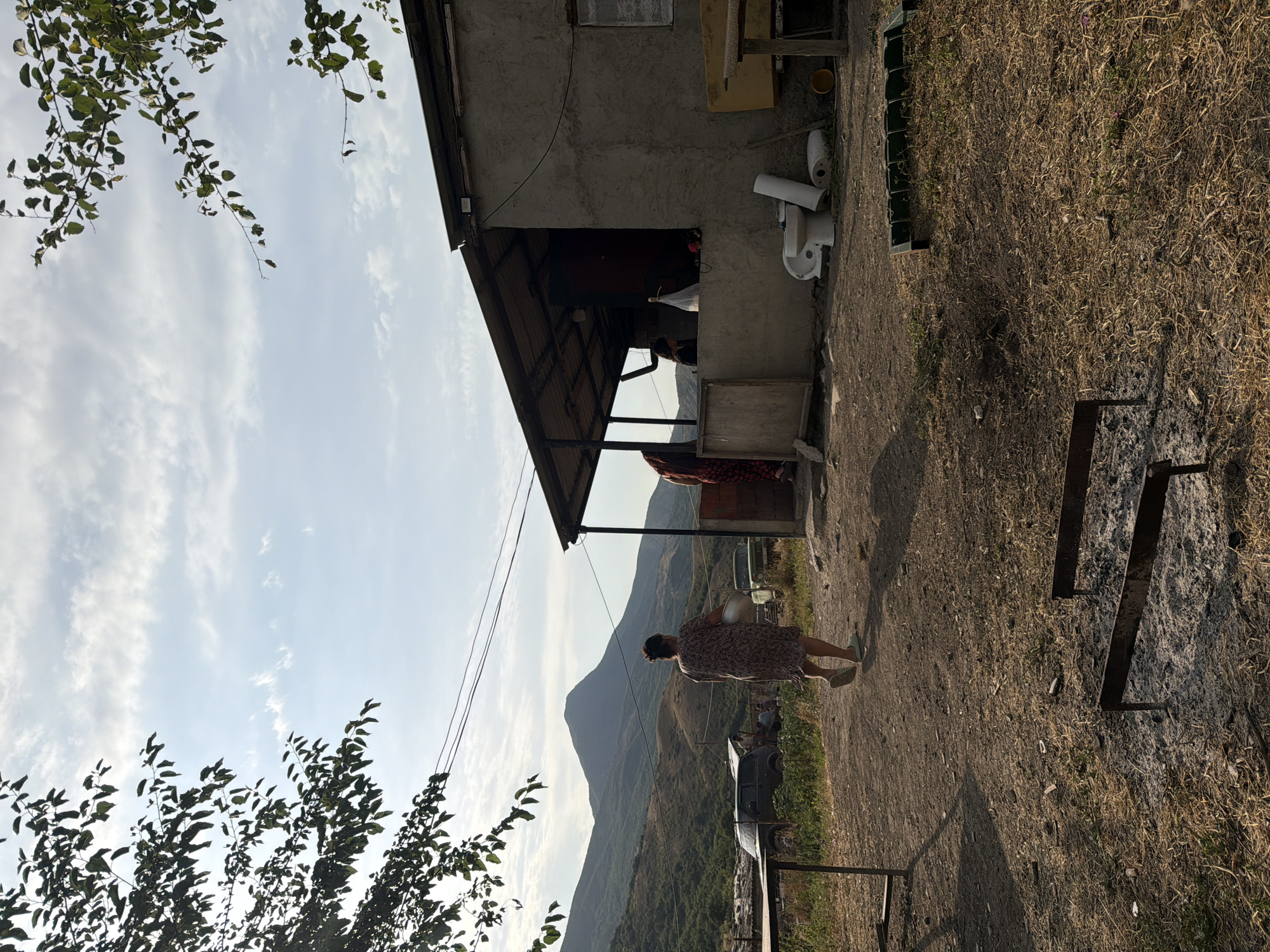 In Köhnə Quşçu, Azerbaijan, an outdoor scene unfolds under a sky dotted with clouds. In the foreground, dry grass and dirt cover the ground, with two metal barbecue grills visible. A table made of wood stands to the left. A woman, seen from behind, walks away from the camera towards a simple, gray, concrete building. She is wearing a patterned dress and light-colored slippers, carrying a large, light-colored bowl. Behind her, a dark SUV is parked, along with another vehicle. In the distance, rolling hills and a prominent mountain peak form the backdrop. The building has a corrugated metal roof supported by posts, and an open porch. Inside the building, partially visible, are people and various objects, including what appears to be a toilet and its tank, along with a yellow bucket. Trees with green leaves frame the upper left and right corners of the image.