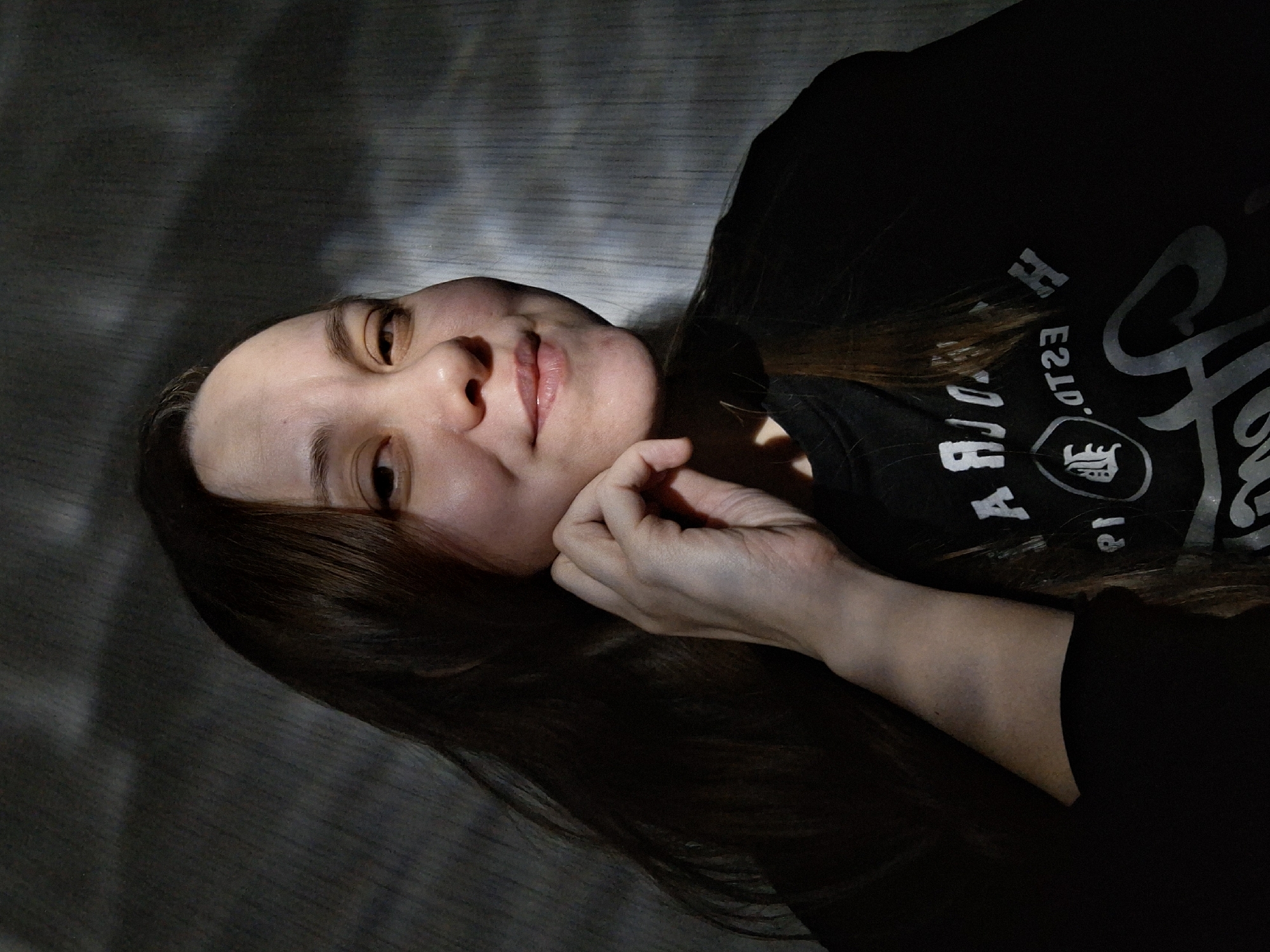 The image is a low-angle portrait of a woman with light skin and long brown hair. She is wearing a black t-shirt with a white graphic design and text. Her right hand is raised near her face. The background appears to be a textured wall with dark shadows. The location, Chaikovsky, Russia, cannot be determined from the image alone.